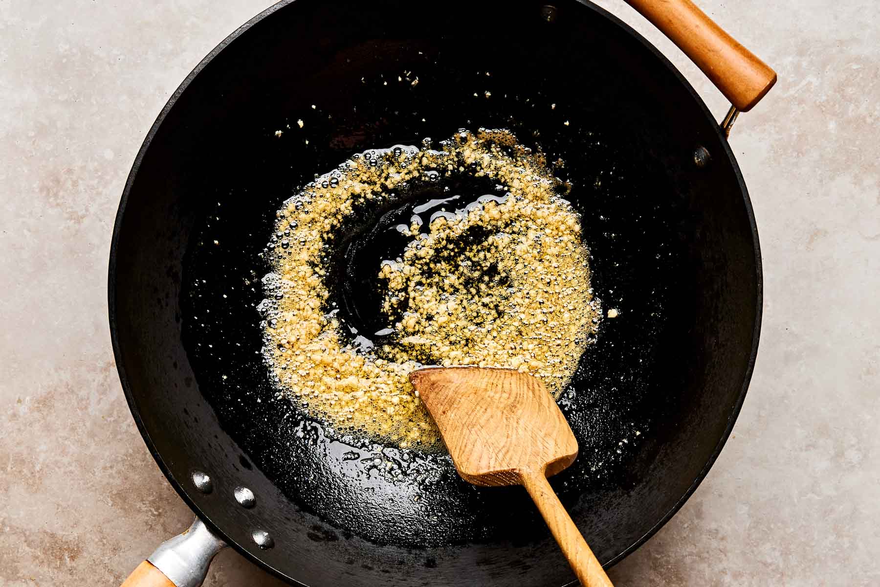Minced garlic being sautéed in oil in a black wok, stirred with a wooden spatula.