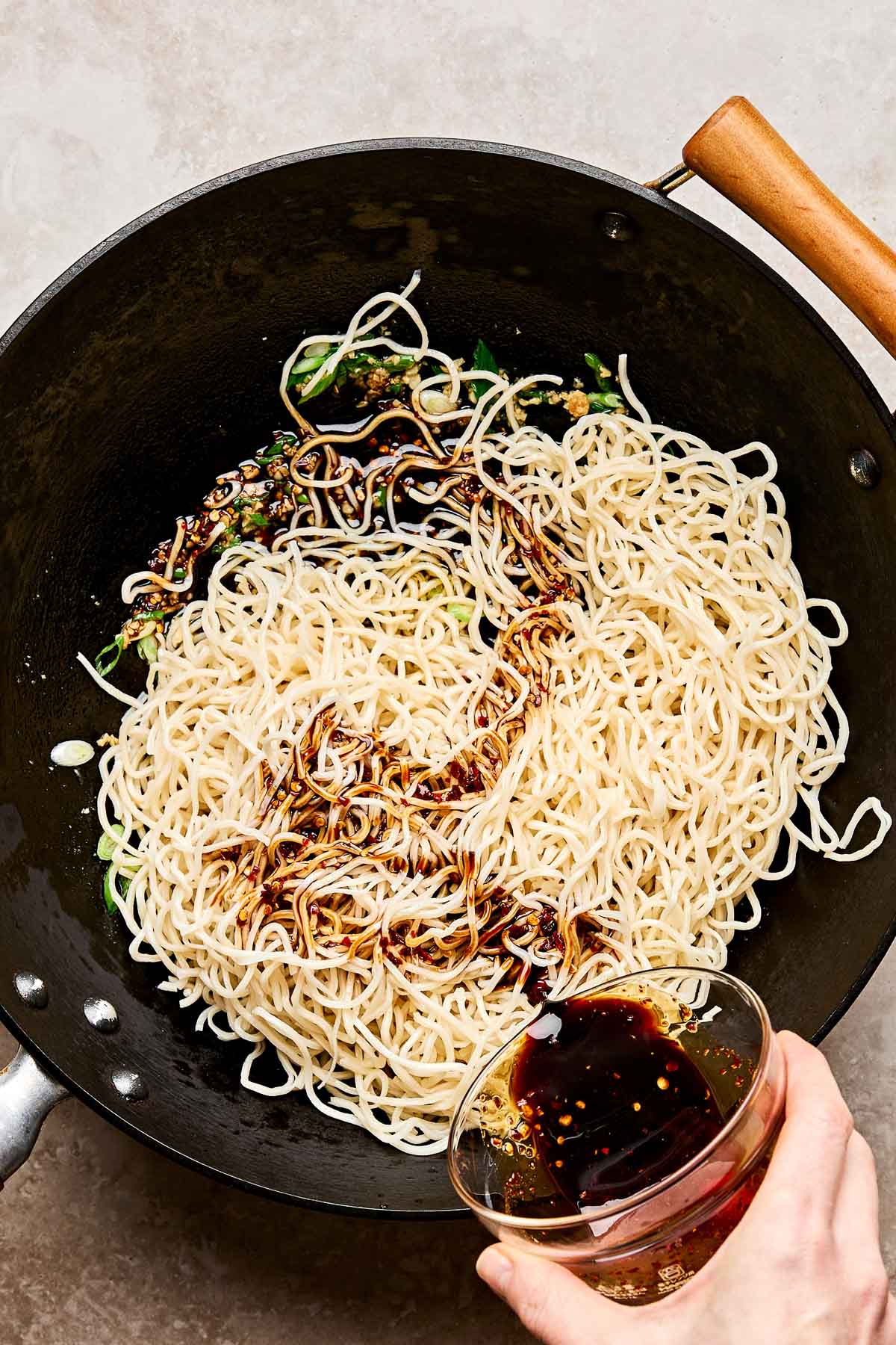 A hand pours a dark soy-based sauce over cooked noodles in a wok, with green onions visible among the noodles. The wok has a wooden handle and is placed on a light countertop.