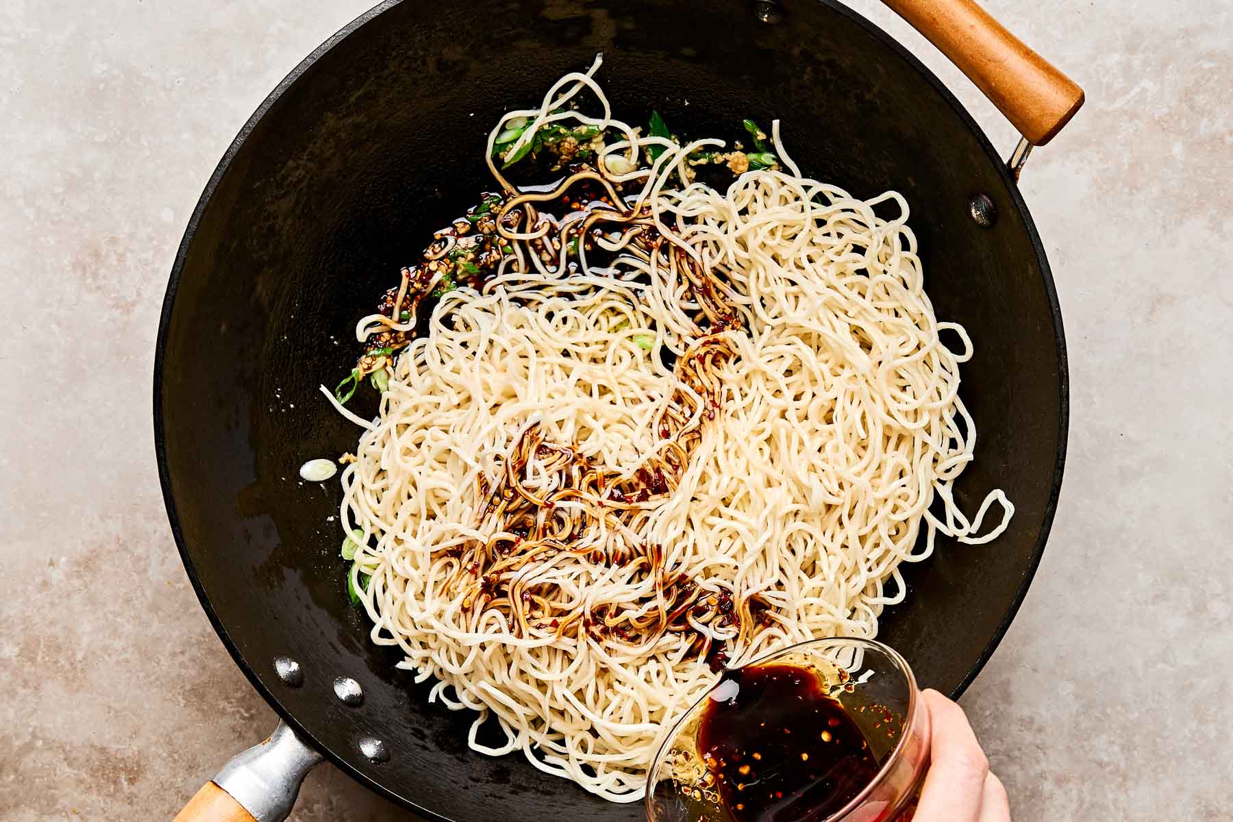 A hand pours dark sauce over cooked noodles in a wok, with green vegetables visible underneath, ready to be mixed together.