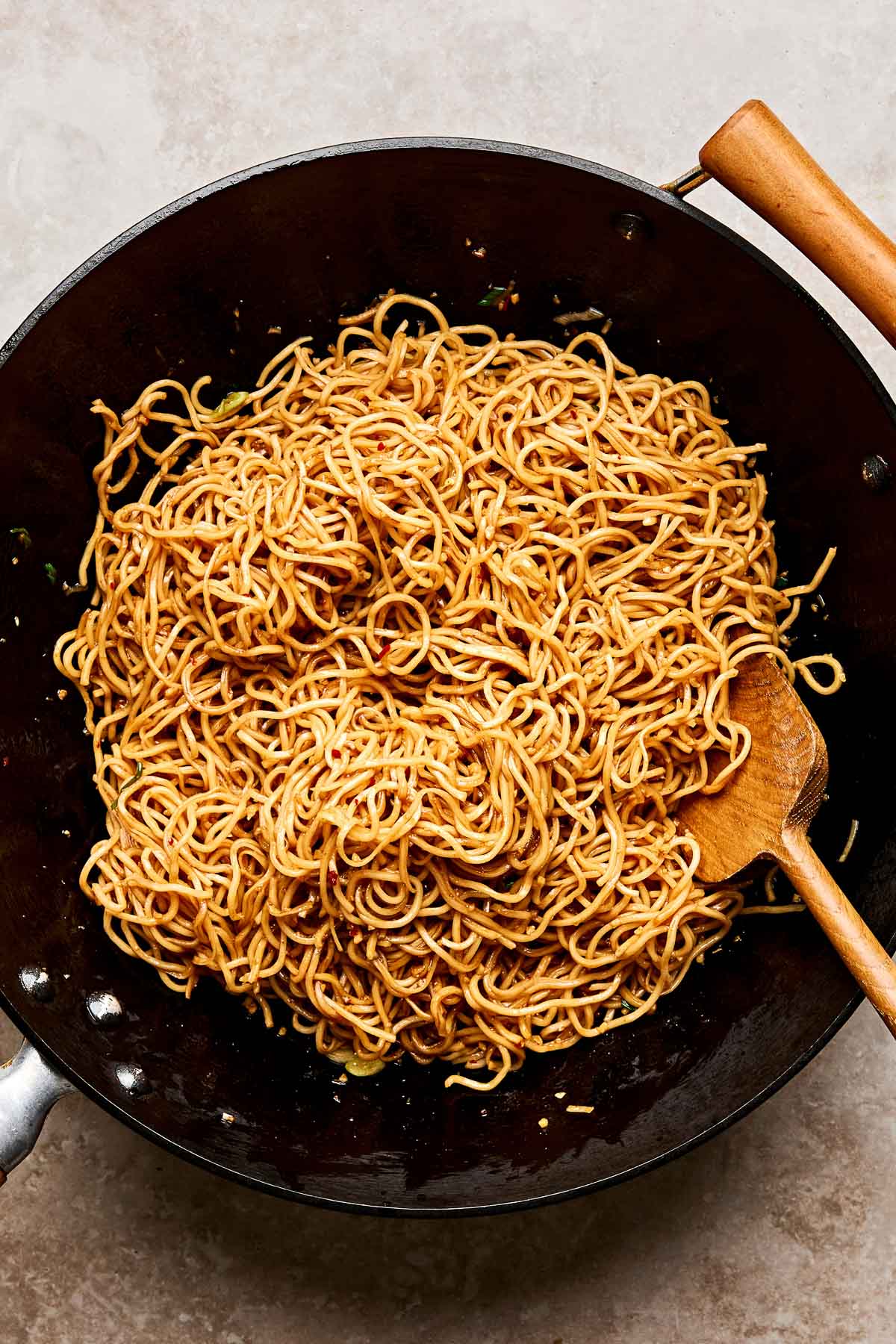 A black wok filled with cooked garlic noodles, mixed with a wooden spoon, placed on a light-colored surface.