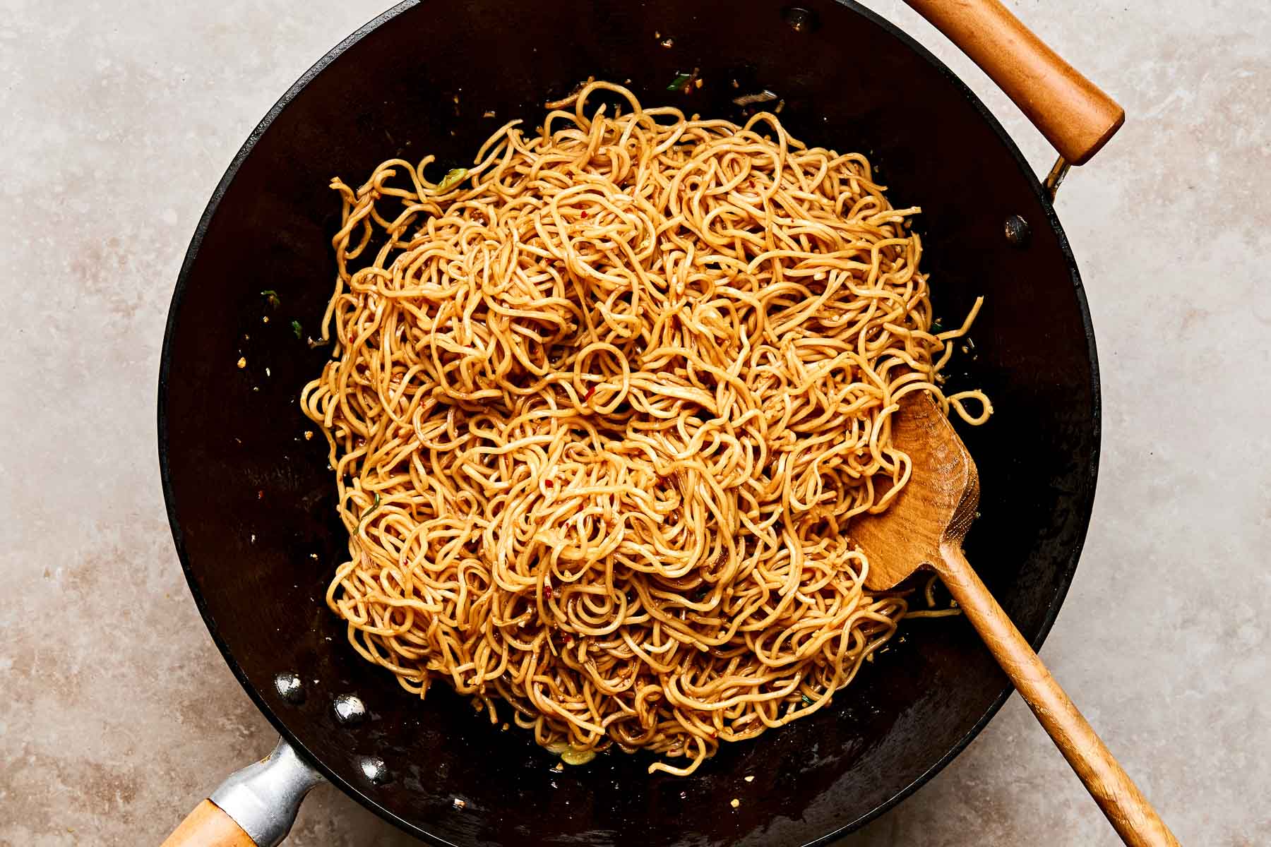 A large black wok filled with cooked soy sauce noodles, mixed and slightly glossy, with a wooden spoon resting inside the wok. The background is a light, textured surface.