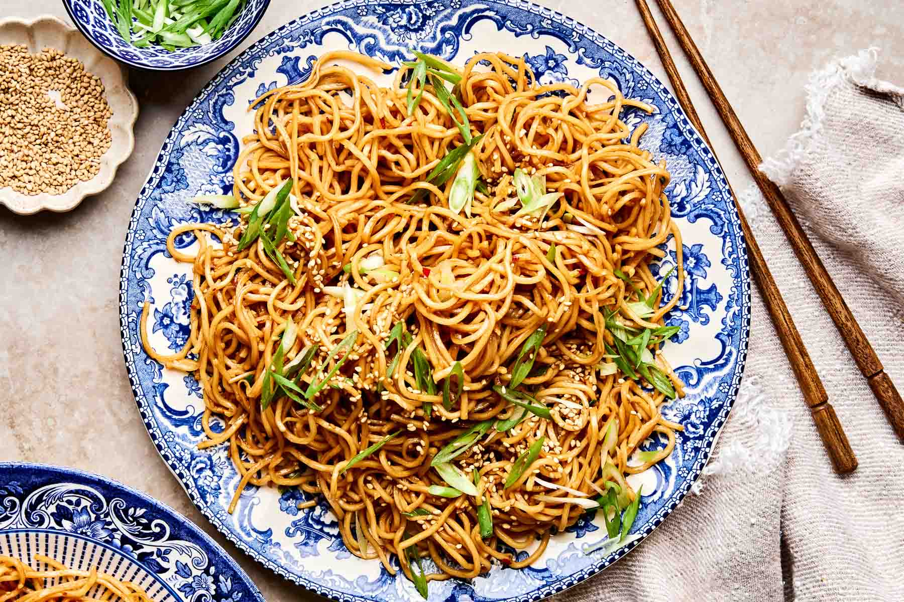 A blue and white plate filled with garlic noodles, garnished with sliced green onions and sesame seeds. Chopsticks, a small bowl of sesame seeds, and a beige napkin are nearby on the table.