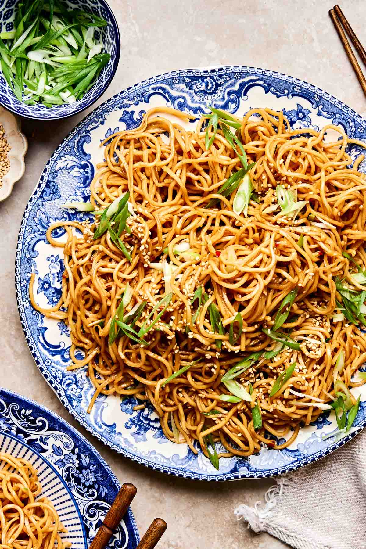 A blue and white patterned plate filled with garlic noodles, garnished with sliced green onions and sesame seeds. Chopsticks and a bowl of sliced green onions are nearby on a beige surface.