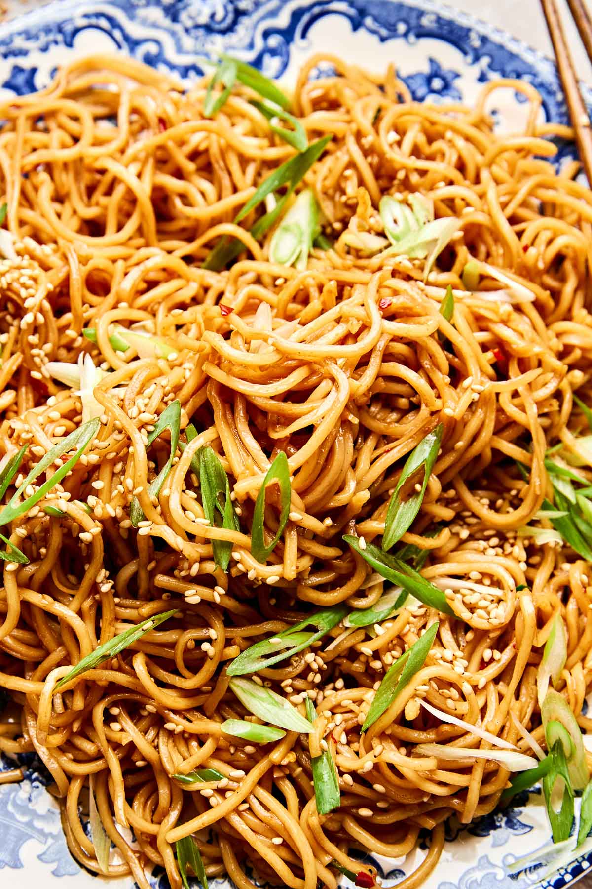 A close-up of a plate of stir-fried garlic noodles garnished with sliced green onions and sesame seeds, served on a decorative blue and white dish with chopsticks.