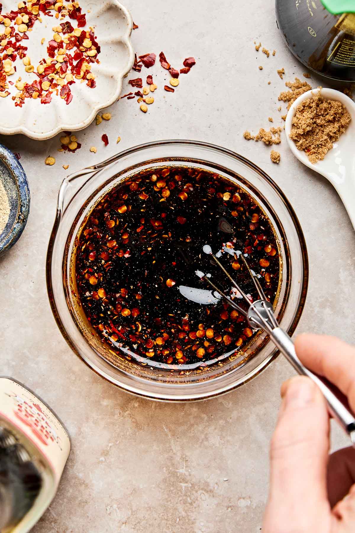 A hand whisking a bowl of dark soy sauce with red pepper flakes on a countertop, surrounded by brown sugar, crushed red pepper, and various seasonings.