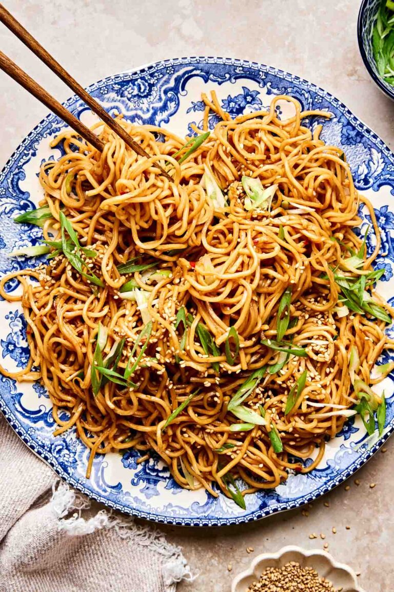 A blue and white plate filled with stir-fried garlic noodles, garnished with sliced green onions and sesame seeds. A pair of wooden chopsticks is lifting some noodles from the plate.