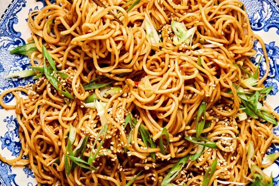 A close-up of stir-fried garlic noodles garnished with sesame seeds and sliced green onions, served on a blue and white patterned plate.