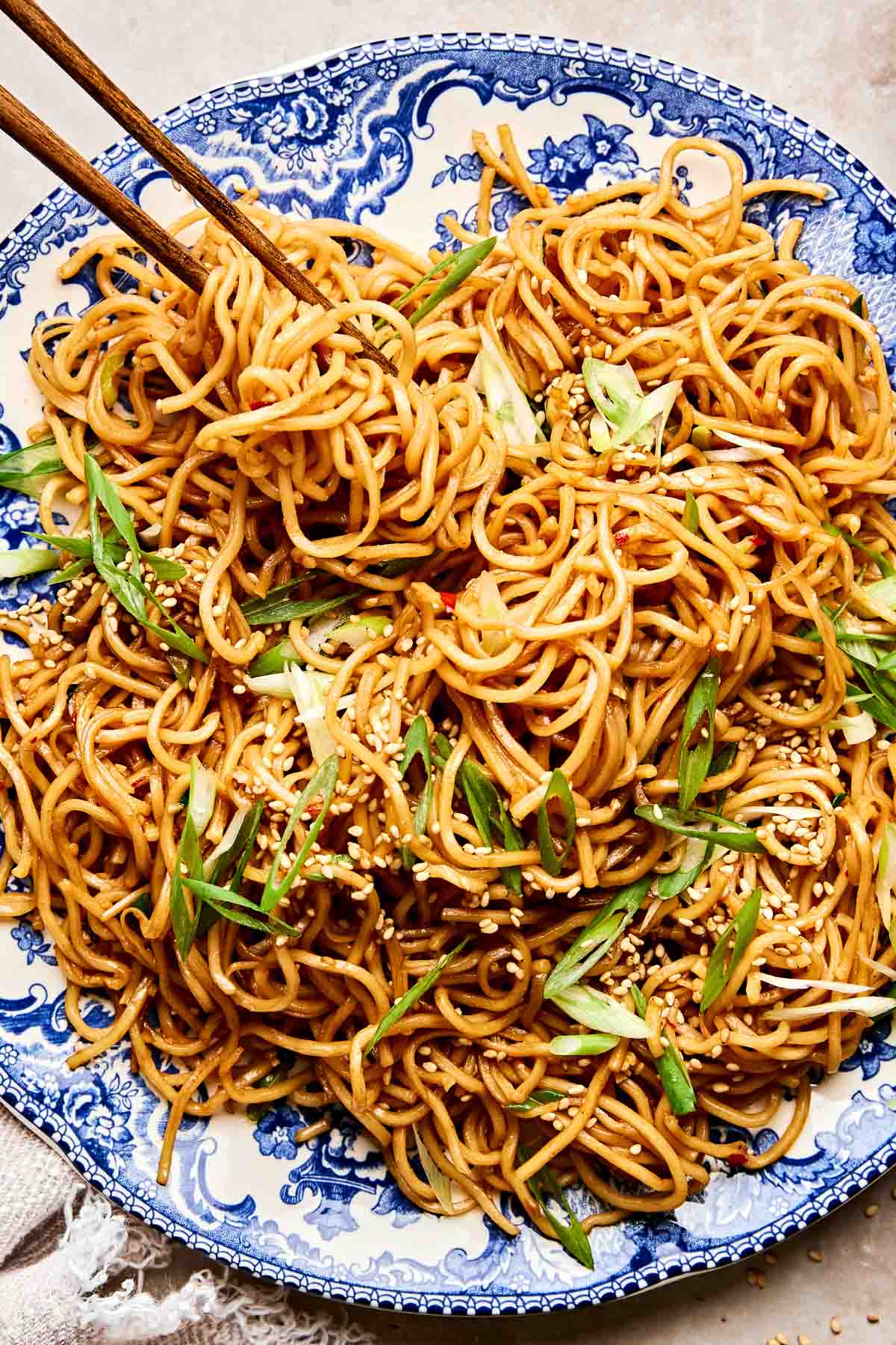 A blue and white plate filled with easy stir-fried garlic noodles, topped with sliced green onions and sesame seeds. A pair of chopsticks rests in the noodles.