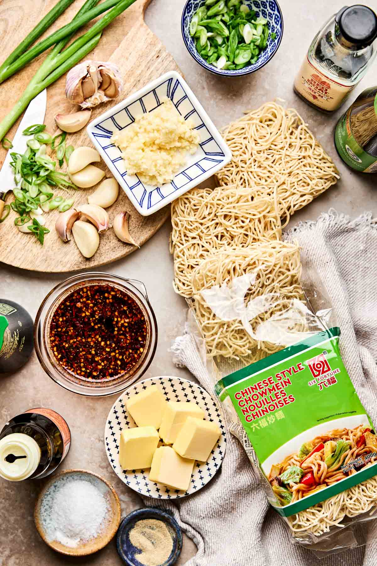 An overhead view of ingredients for a noodle dish including uncooked Chinese style noodles, butter, chopped scallions, garlic, ginger, chili oil, soy sauce, and seasonings arranged on a kitchen surface.