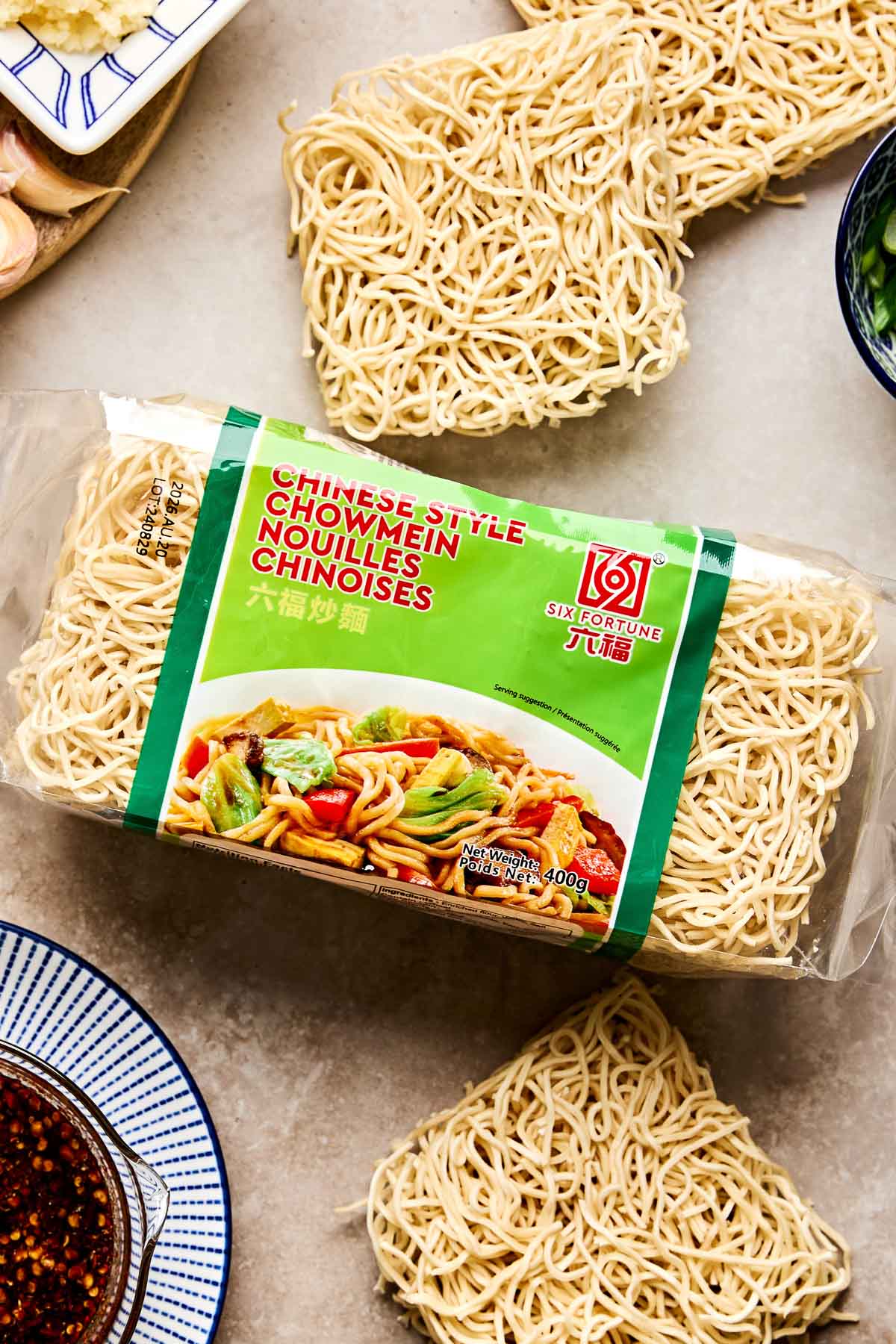 A package of Six Fortune Chinese Style Chow Mein noodles sits on a counter, surrounded by uncooked noodles, a bowl of sauce, and some vegetables. The package shows an image of stir-fried noodles with vegetables.