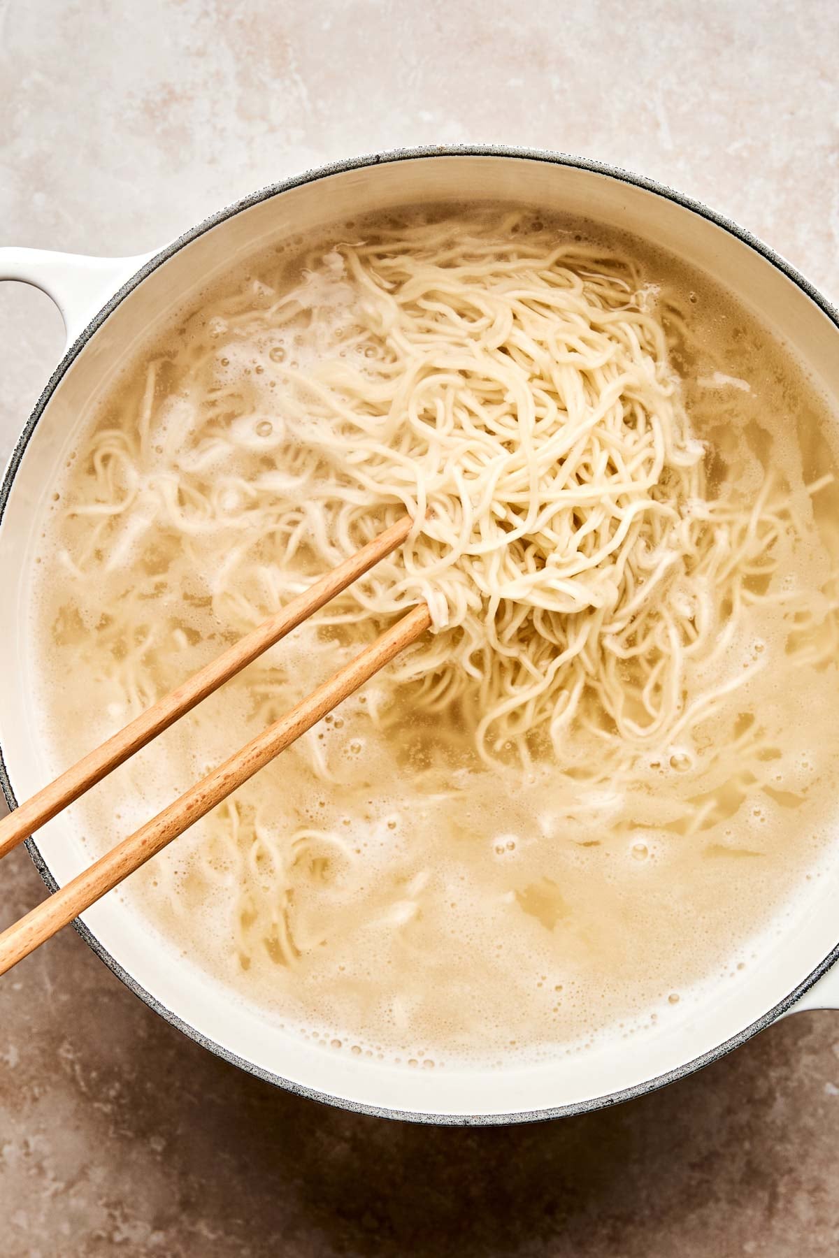 A close-up of a pot filled with boiling water and chow mein noodles being lifted with wooden chopsticks. The noodles are partially submerged, and steam rises from the pot.