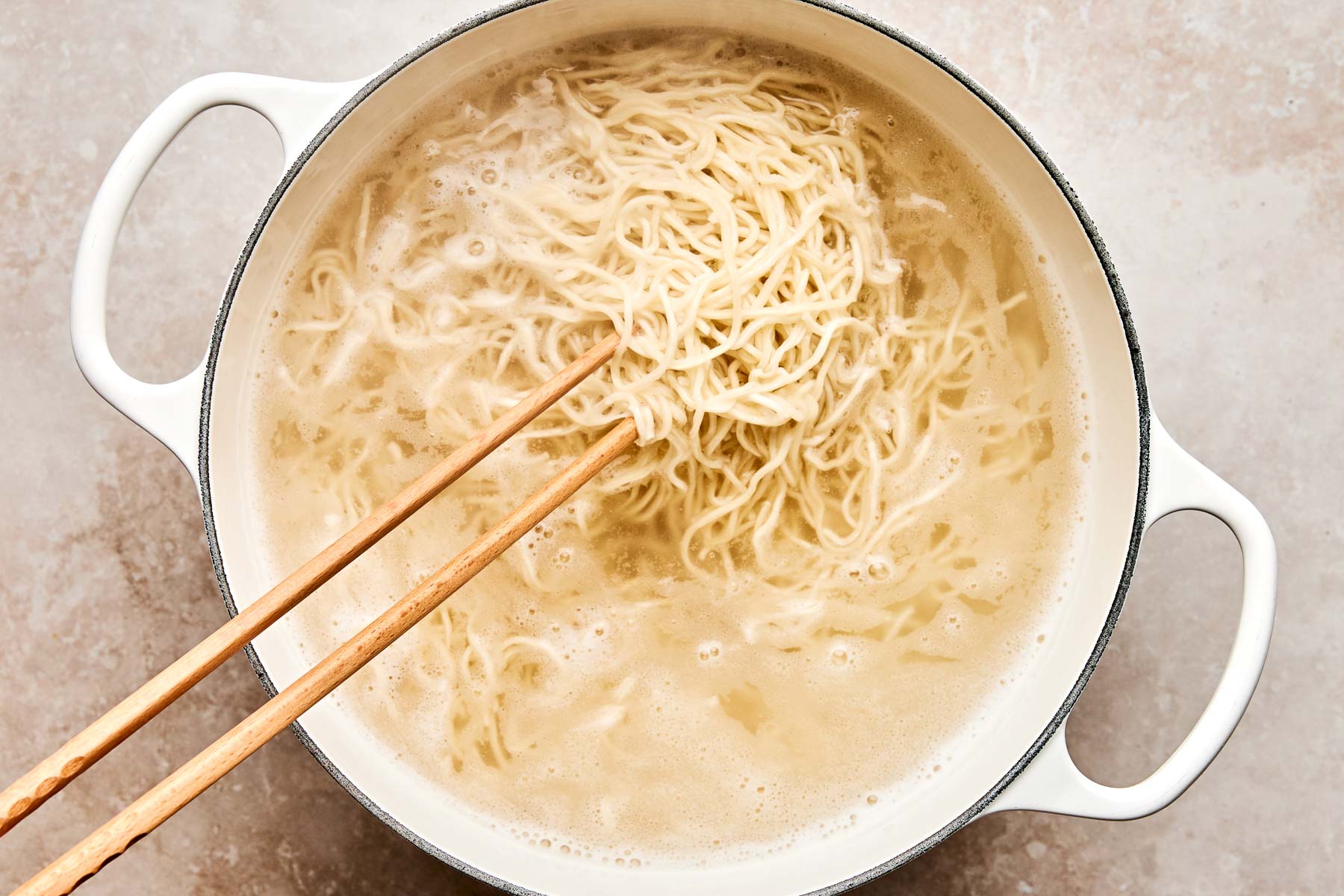 A pot of boiling water with noodles being stirred using wooden chopsticks, seen from above on a light-colored surface.