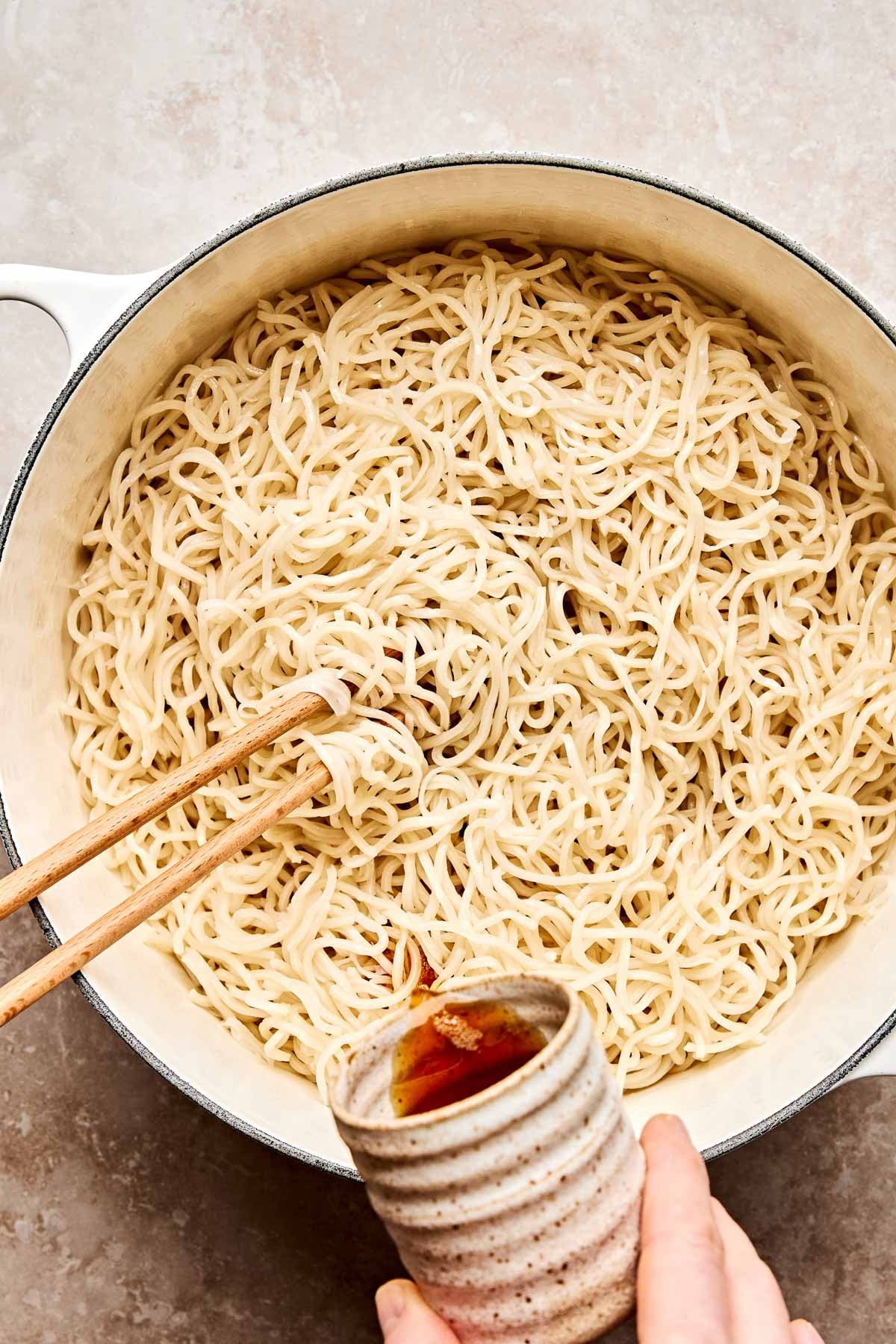 A large white pot filled with cooked noodles being stirred with wooden chopsticks, while a hand pours sauce from a small ceramic cup over the noodles.
