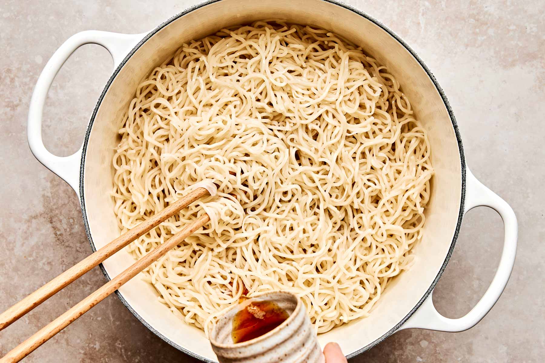 A large white pot filled with cooked noodles, with wooden chopsticks stirring them, while a hand pours sauce from a small ceramic bowl into the pot.