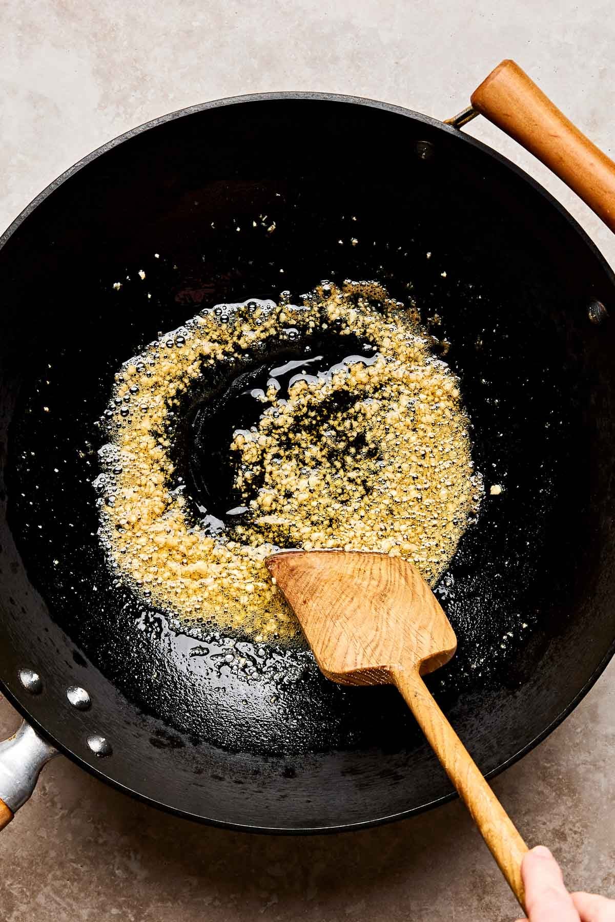 A wooden spatula stirs minced garlic sautéing in oil inside a black wok on a light countertop.