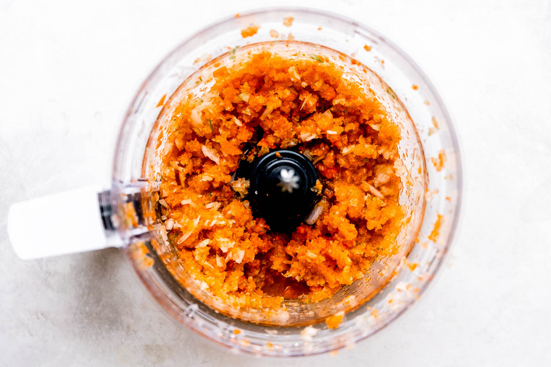 A top-down view of a food processor containing finely chopped orange vegetables and onions, creating a coarse mixture on a light countertop.