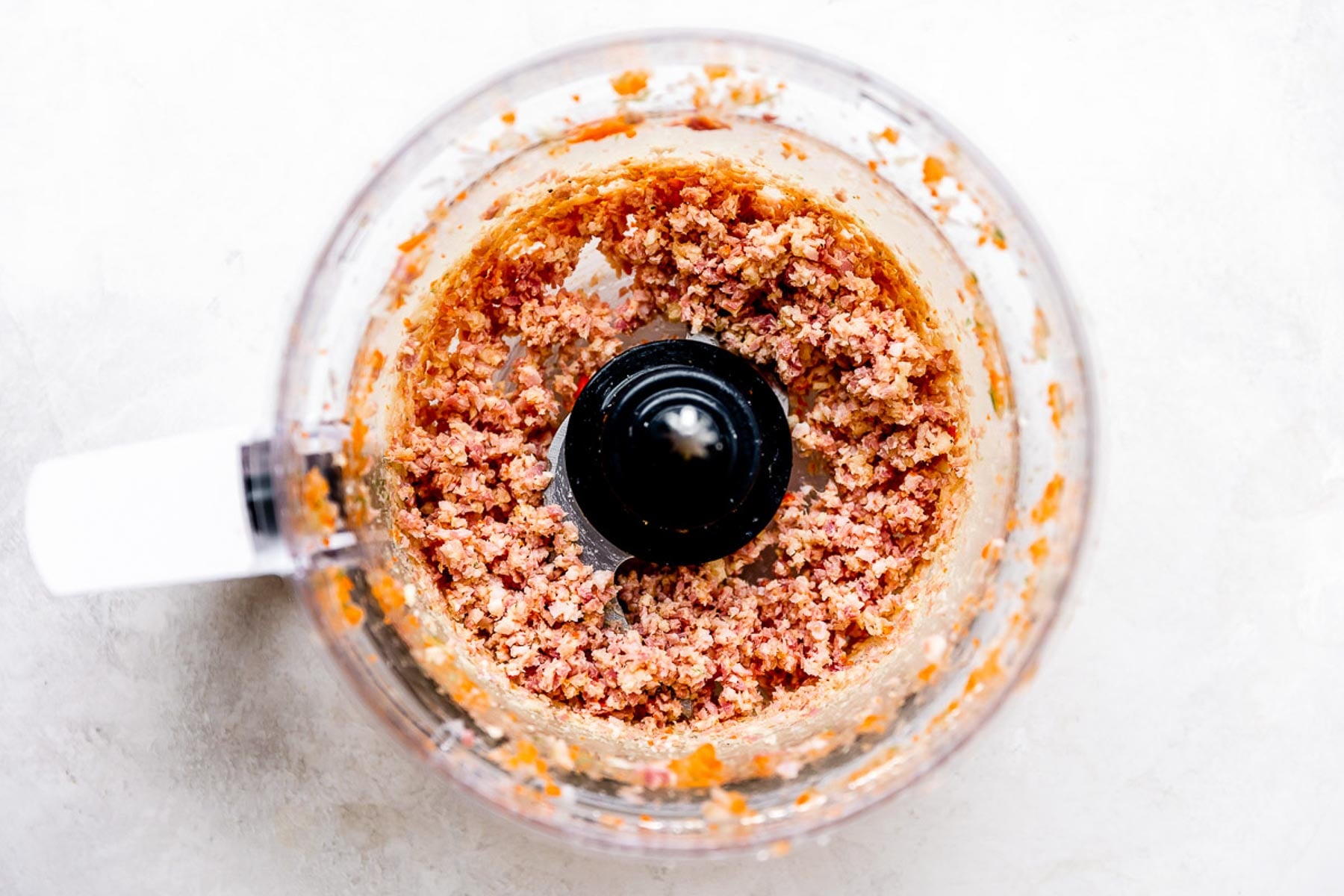 A food processor bowl containing finely chopped or blended ingredients with a reddish-orange color, viewed from above on a light surface.