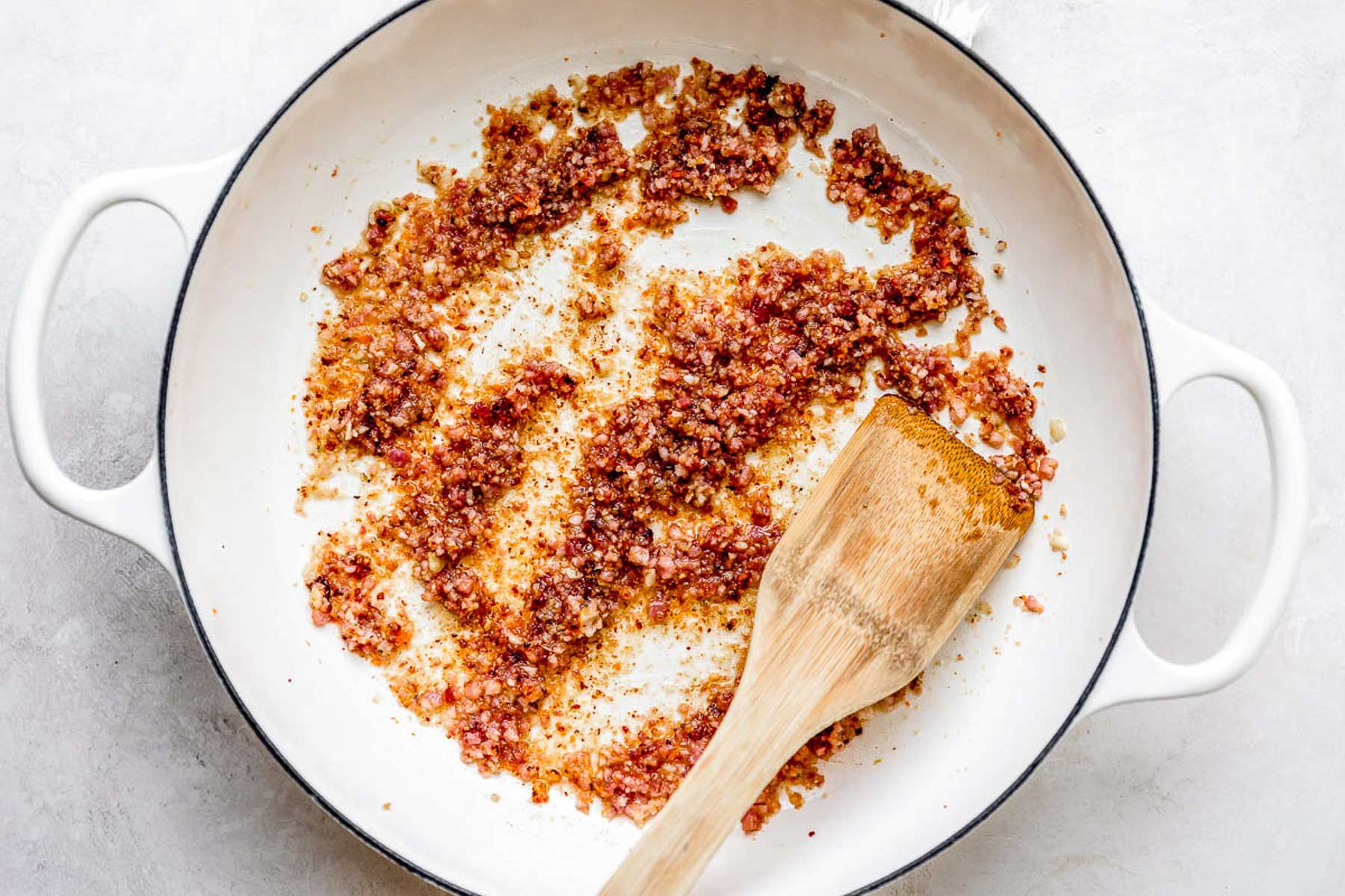 A white skillet containing finely chopped, sautéed bacon bits, with a wooden spatula resting inside the pan. The pan is set on a light-colored surface.