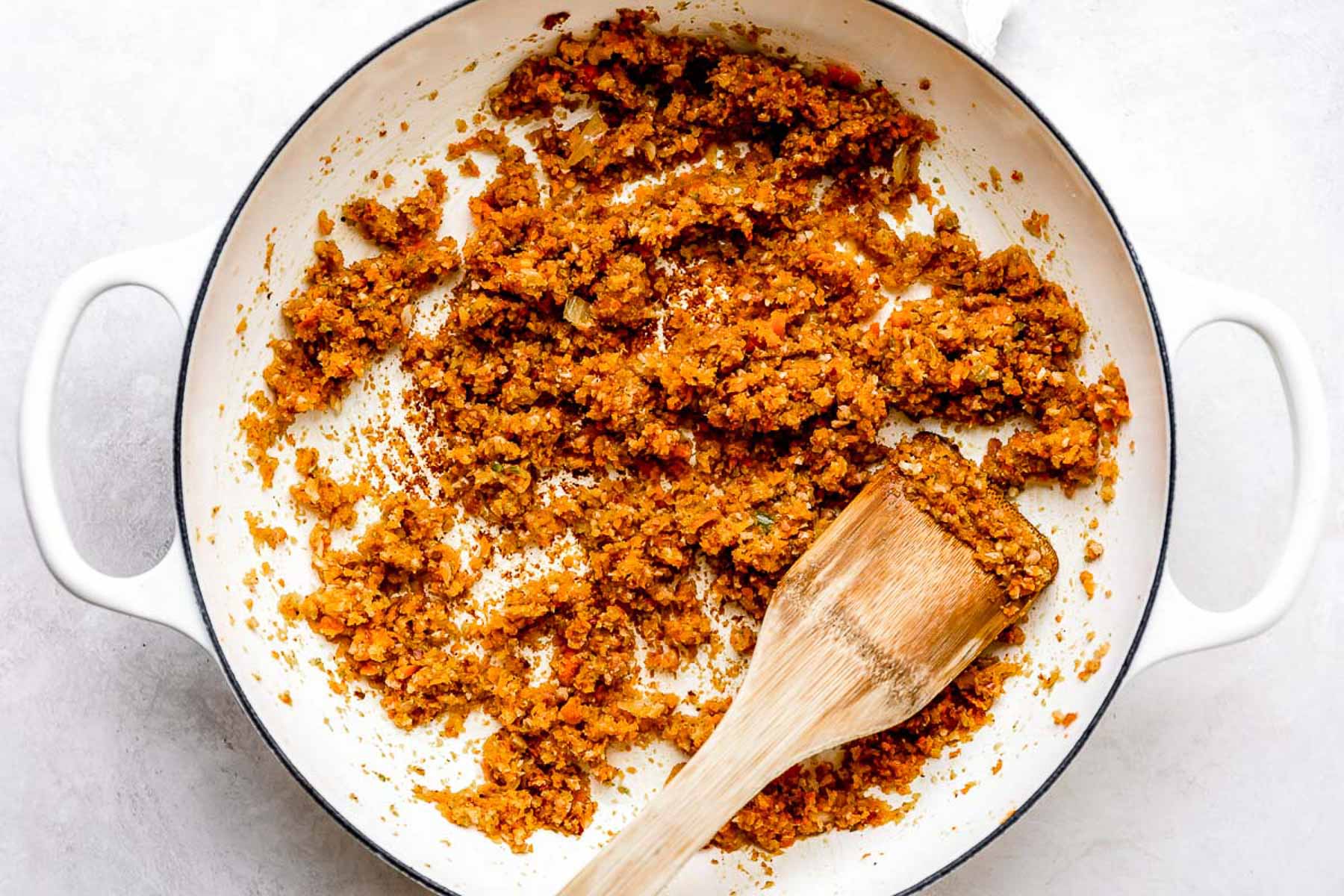 A white pot with browned, finely chopped vegetables being sautéed, stirred with a wooden spatula. The mixture looks like classic mirepoix or sofrito, used as a flavor base for cooking.