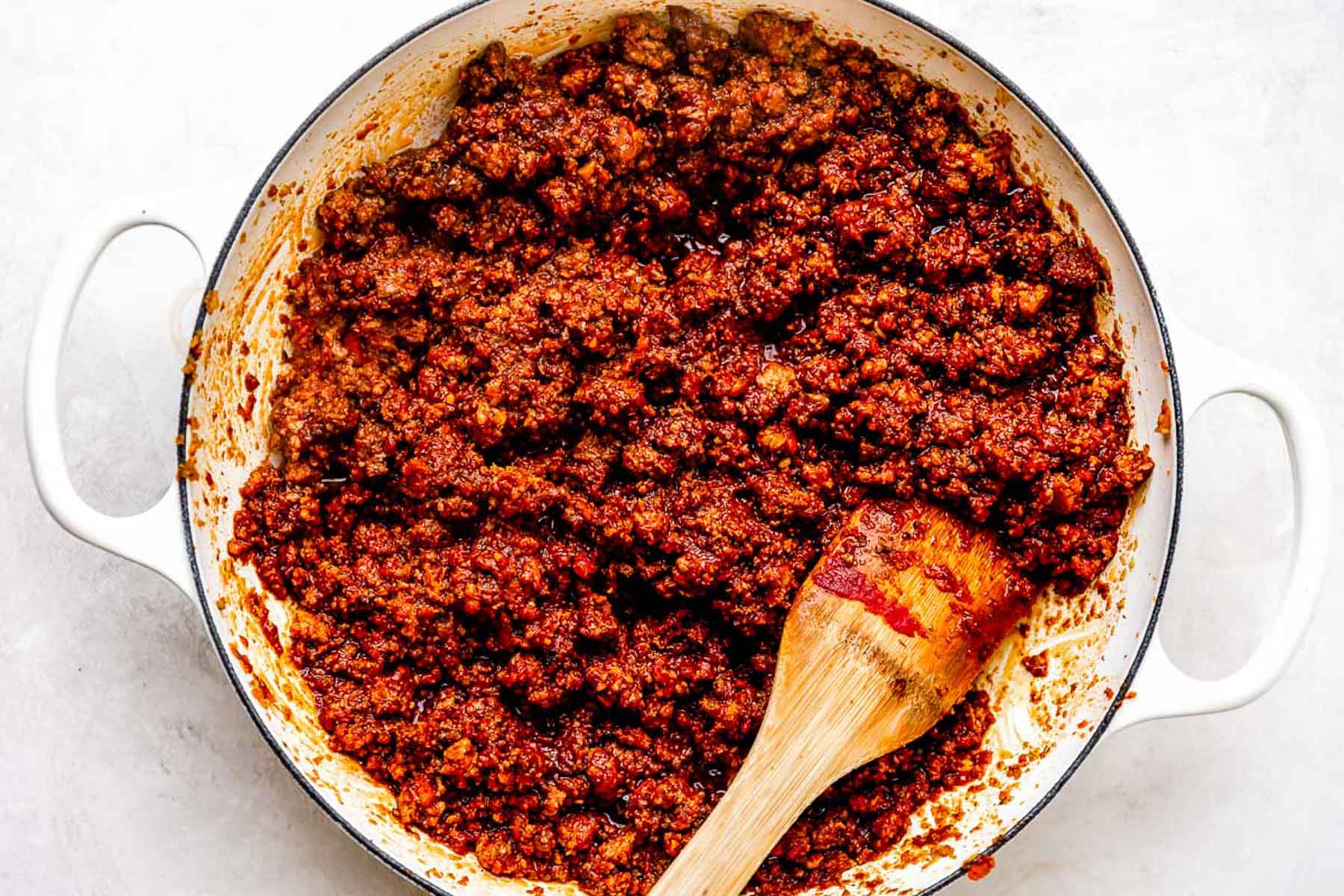 A white pot filled with cooked ground beef taco meat in a rich, reddish sauce, with a wooden spoon resting inside.