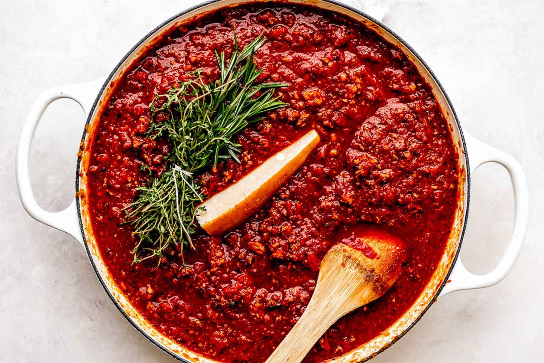 A pot of rich red tomato sauce with ground meat, topped with fresh rosemary and thyme, and a wedge of Parmesan cheese, stirred with a wooden spoon.