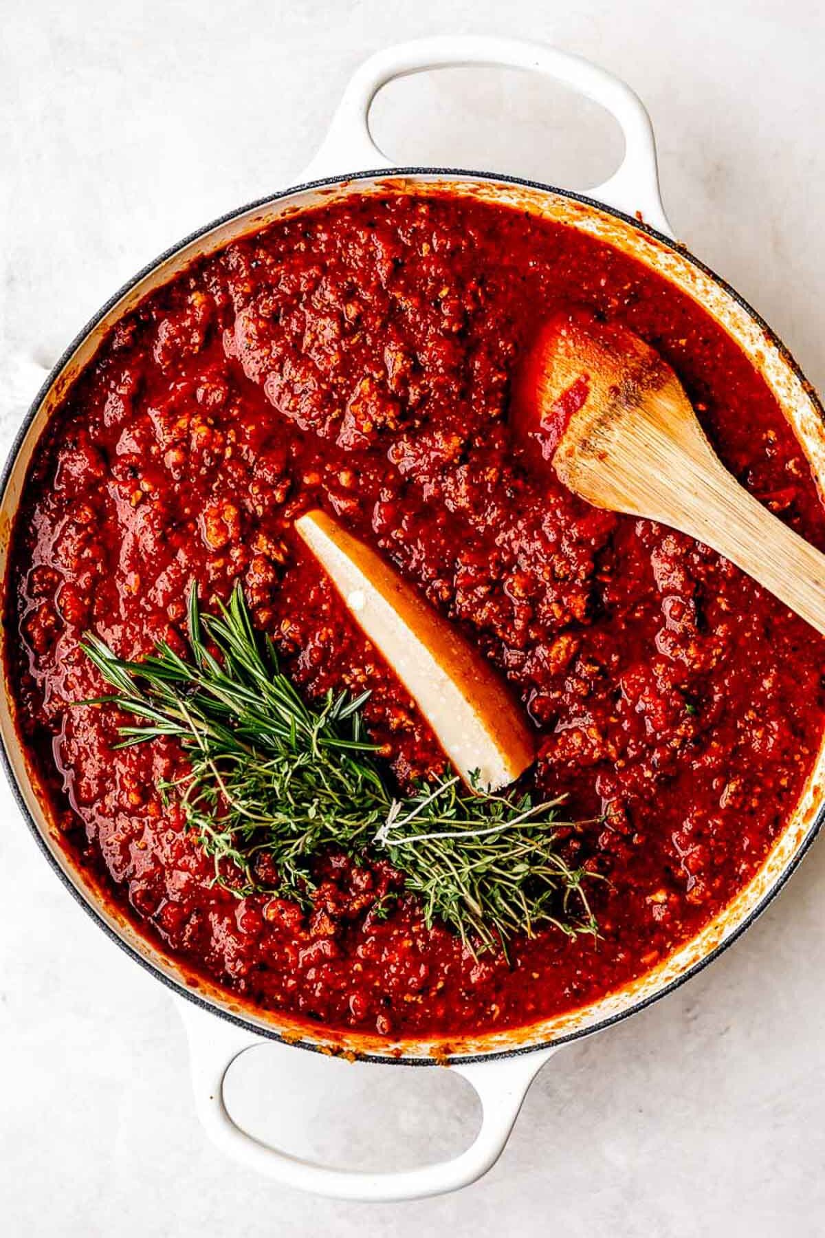 A pot of rich red tomato sauce with visible ground meat, a cluster of fresh herbs, and a wedge of Parmesan cheese on top. A wooden spoon rests in the sauce. The pot is white and set on a light surface.