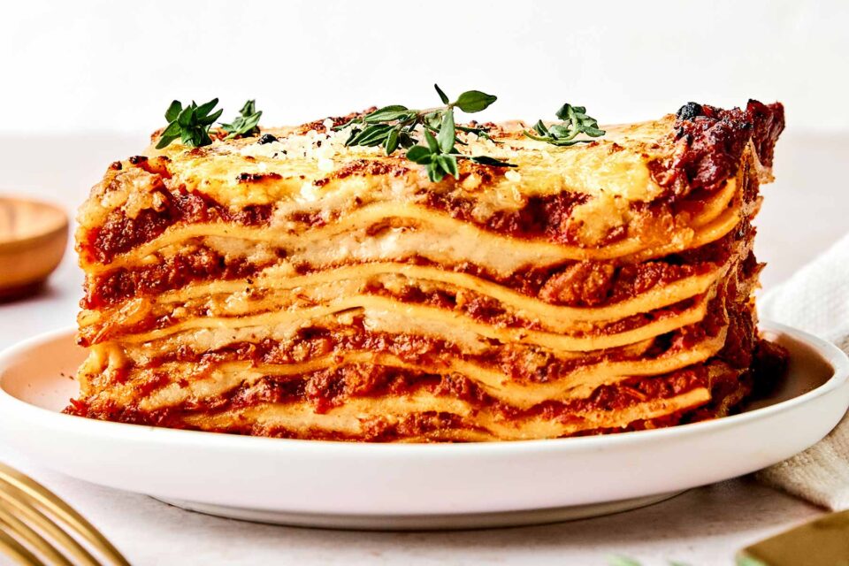 A close-up of a thick slice of lasagna on a white plate, showing multiple layers of pasta, tomato sauce, cheese, and herbs, garnished with fresh thyme on top.
