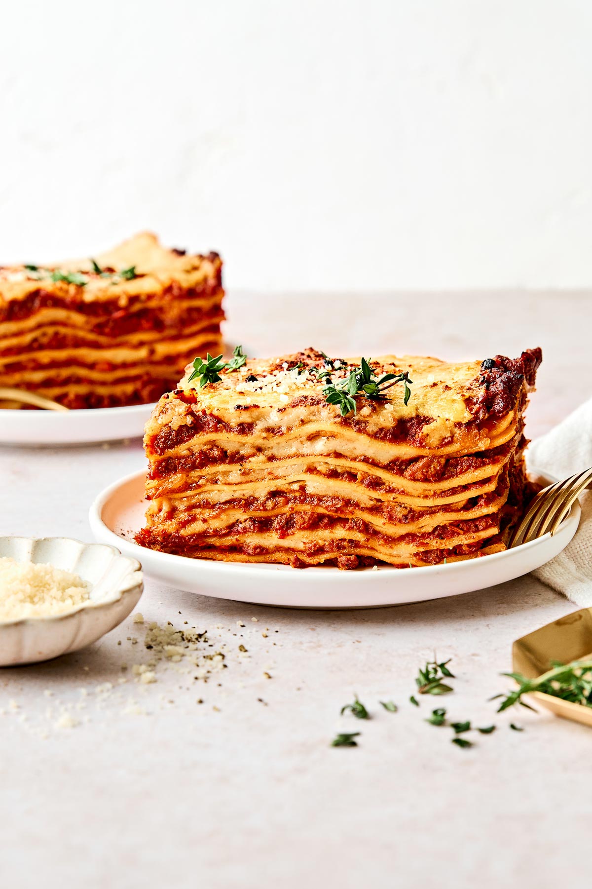 A large slice of layered lasagna topped with herbs is served on a white plate, next to a fork and a small dish of grated cheese. Another slice of lasagna is in the background.