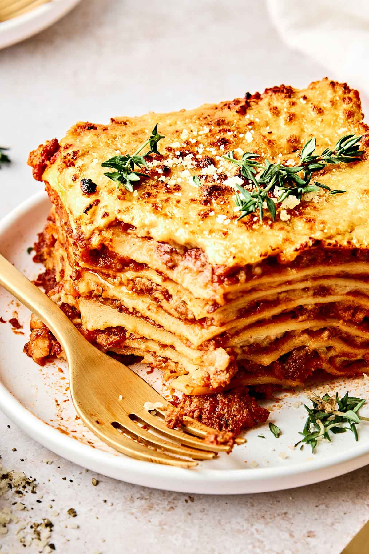 A close-up of a slice of lasagna on a white plate, garnished with fresh herbs and grated cheese. A gold fork lies next to the lasagna on the plate.