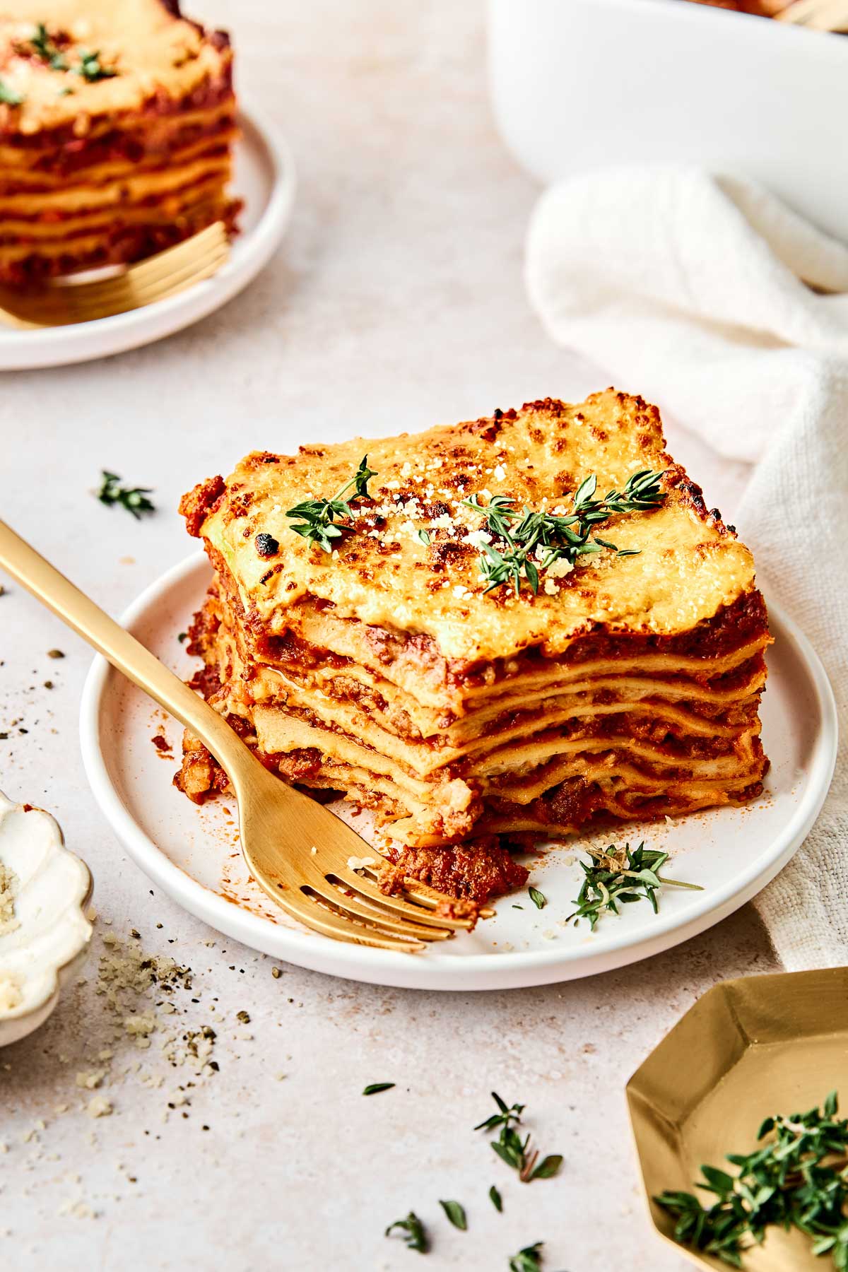 A slice of lasagna with layers of pasta, sauce, and cheese is served on a white plate, garnished with fresh herbs. A gold fork rests beside the lasagna, and another plate and baking dish are partially visible in the background.