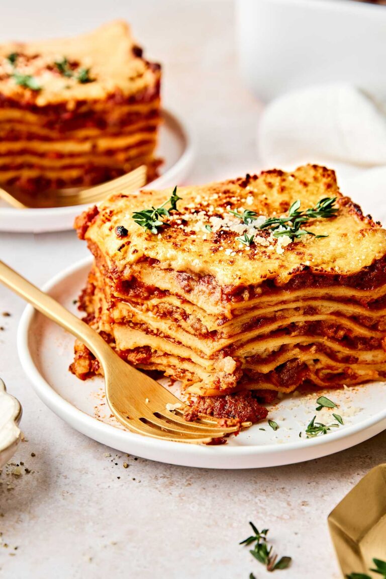 A slice of cheesy lasagna with visible layers of pasta, sauce, and cheese, garnished with fresh herbs, served on a white plate with a gold fork. Another slice is in the background.