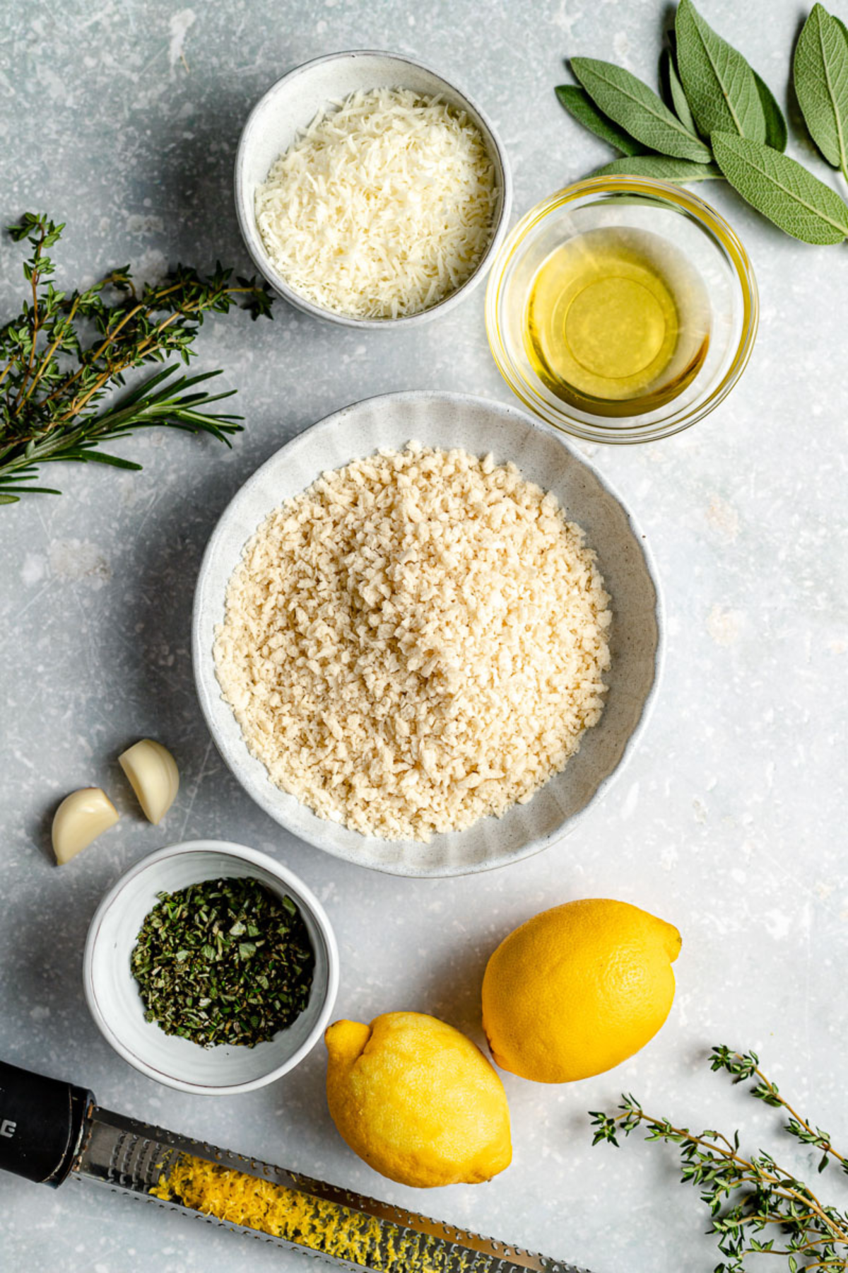 A bowl of breadcrumbs surrounded by a bowl of grated cheese, a bowl of chopped herbs, fresh herbs, two lemons, two garlic cloves, a bowl of olive oil, and a zester on a light gray surface.