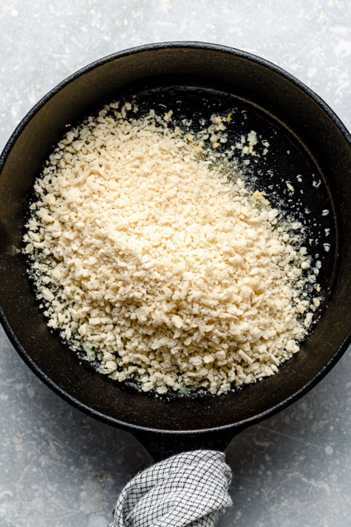 A black skillet filled with a mound of light-colored breadcrumbs, resting on a gray textured surface with a checkered cloth wrapped around the handle.
