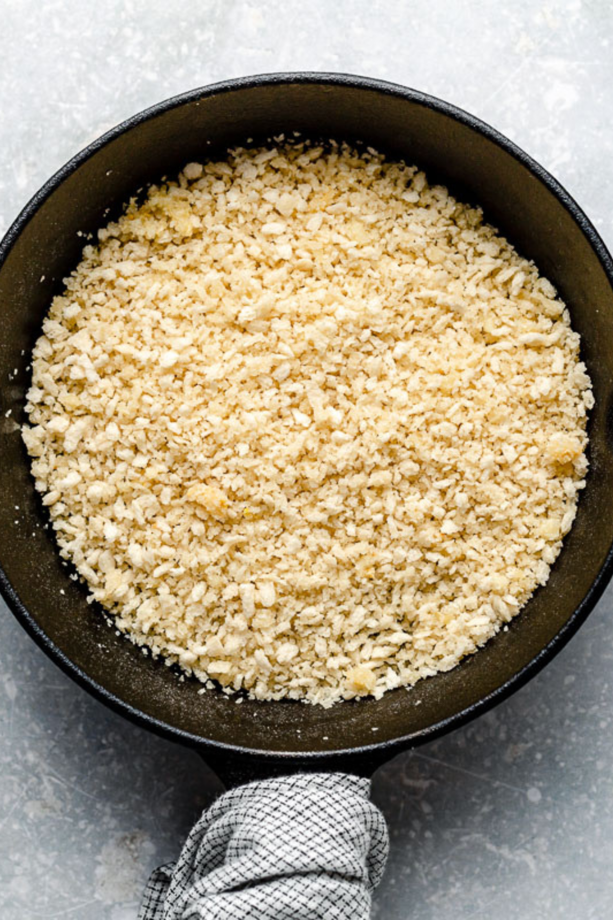 A black pan filled with toasted breadcrumbs sits on a gray surface, with a checkered cloth wrapped around its handle.