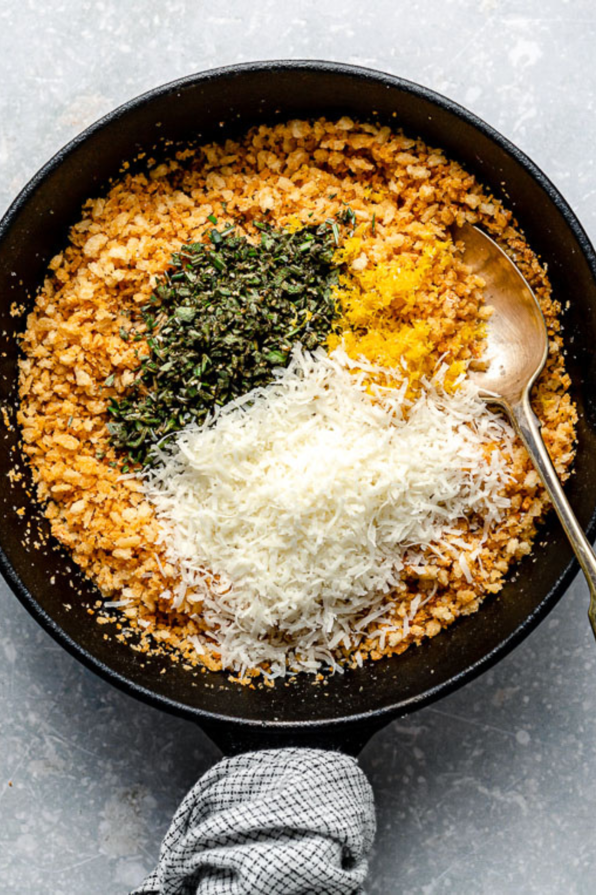 A black skillet filled with breadcrumbs, grated cheese, lemon zest, and chopped herbs, with a gold spoon, sits on a gray surface. A gray kitchen towel is wrapped around the handle.