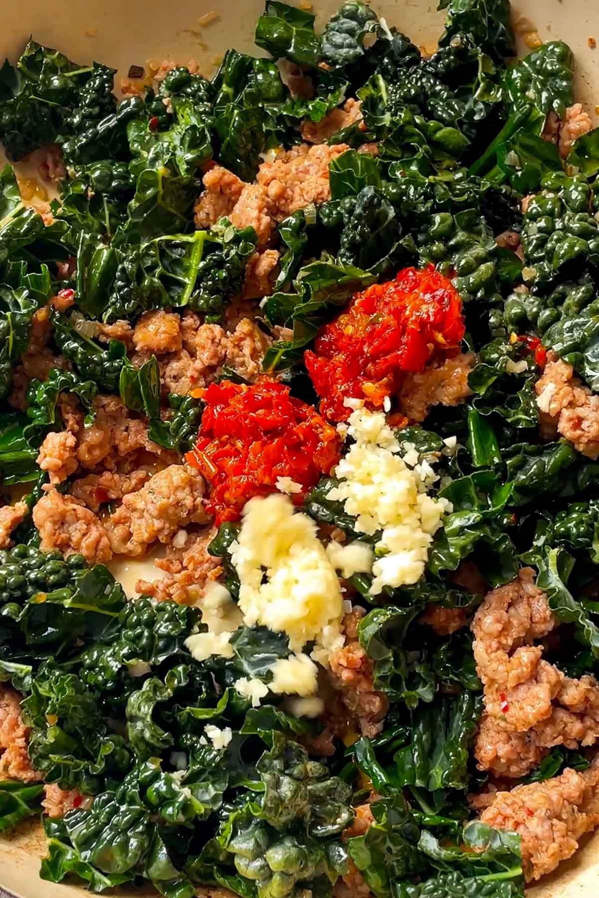 Close-up of a skillet filled with sautéed kale, crumbled sausage, crushed red tomatoes, and minced garlic, creating a colorful and hearty mixture.
