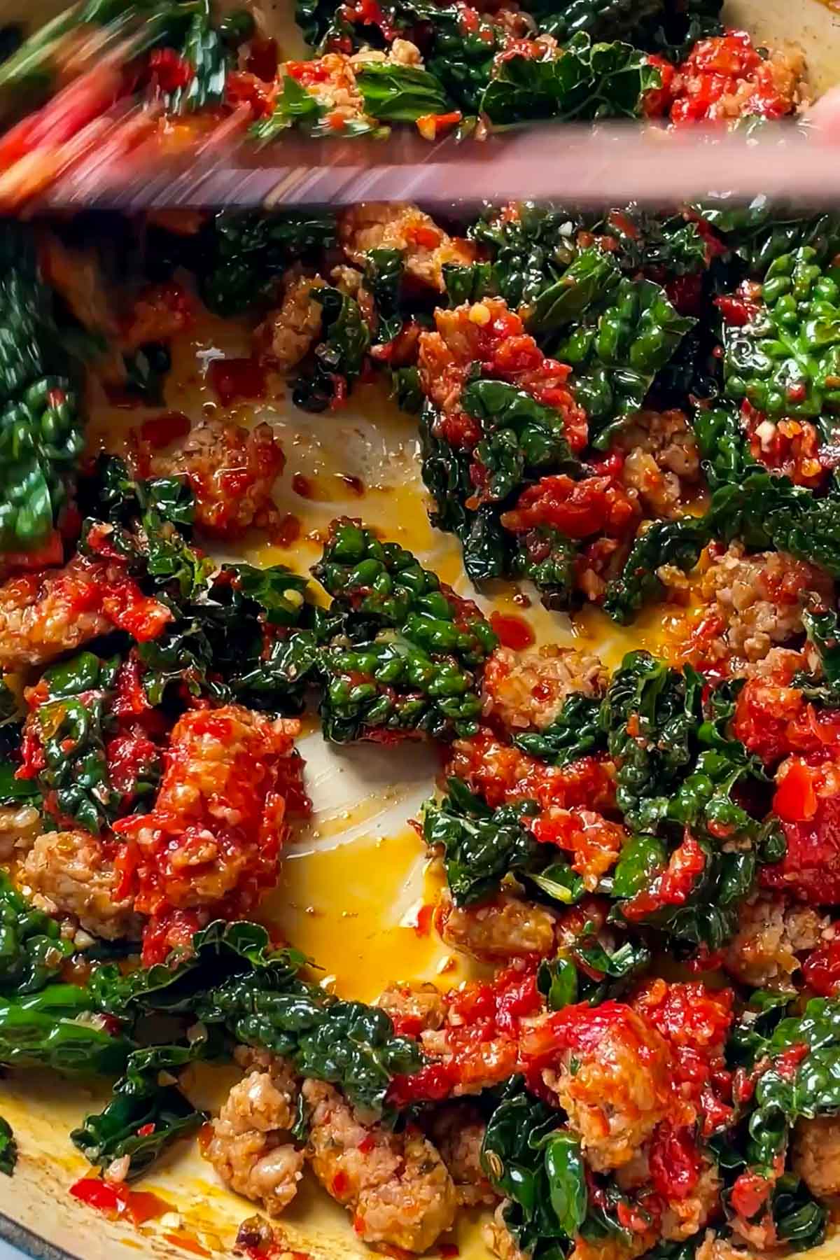 Close-up of a skillet with cooked sausage, wilted dark green kale, and chunky tomato sauce being stirred together, showing vibrant colors and textures.