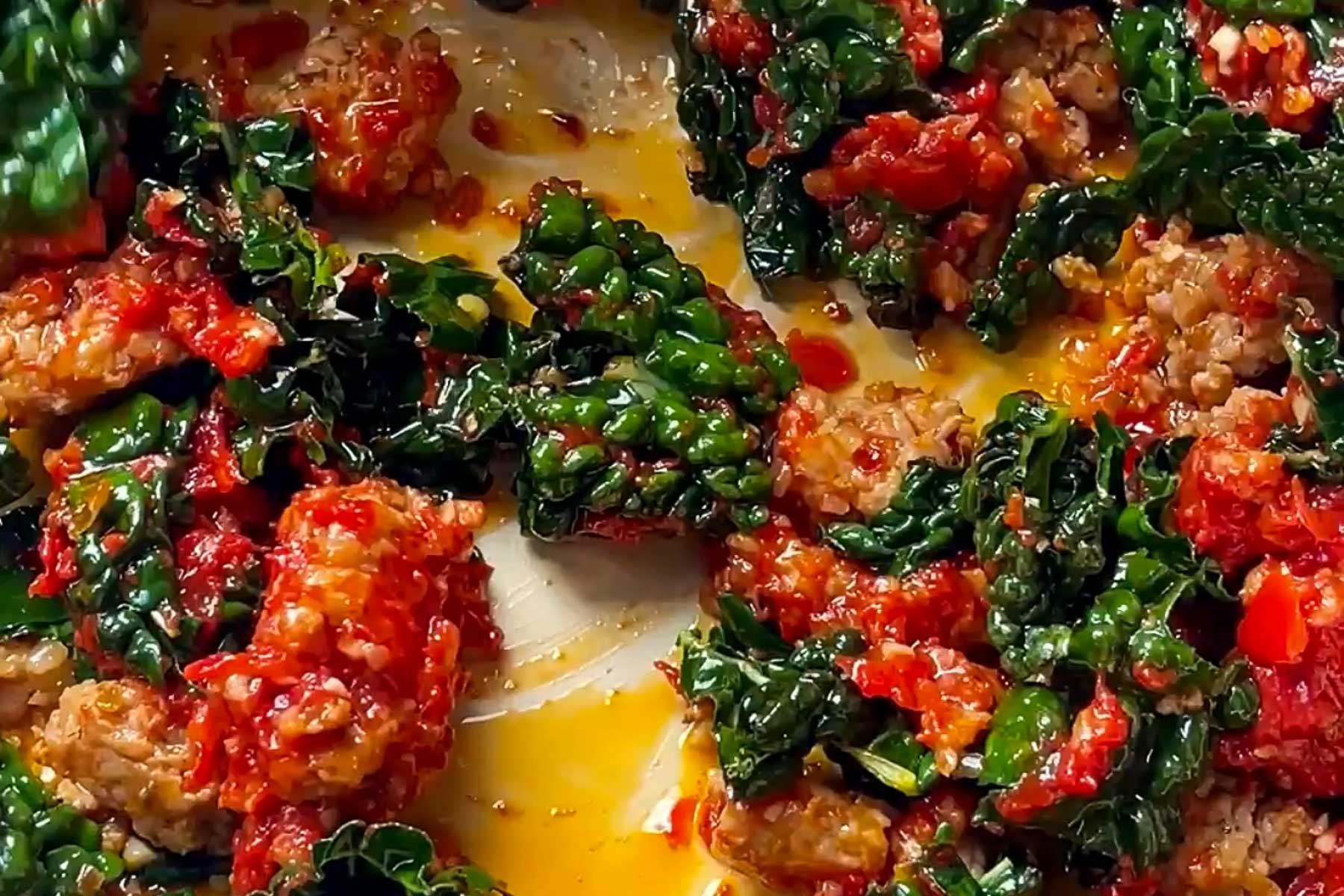 Chunks of sausage, wilted kale, and pieces of tomato are cooked together in a pan, surrounded by a flavorful orange-colored sauce.