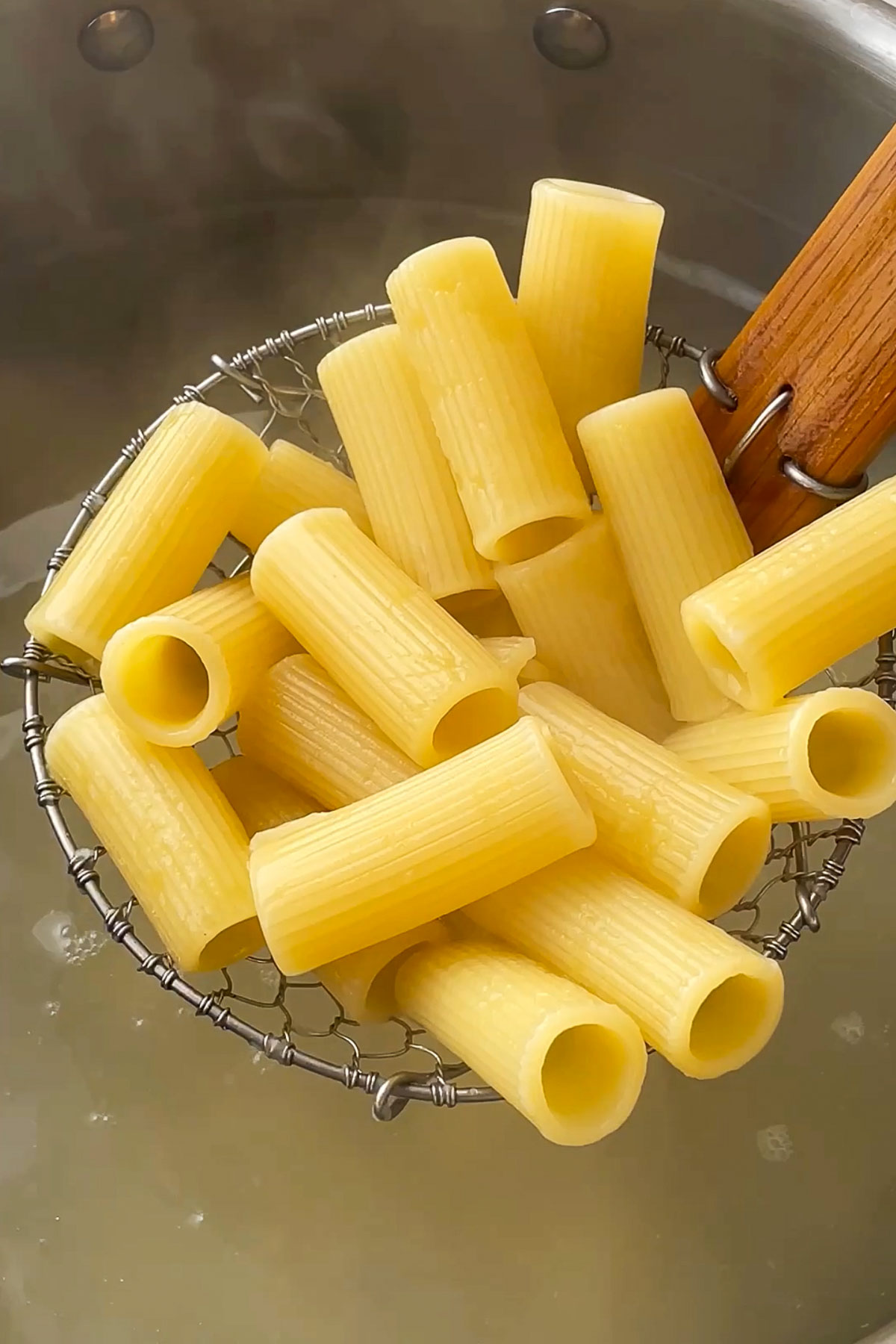 A wire strainer holds cooked rigatoni pasta above a pot of boiling water, with a wooden handle visible on the right.