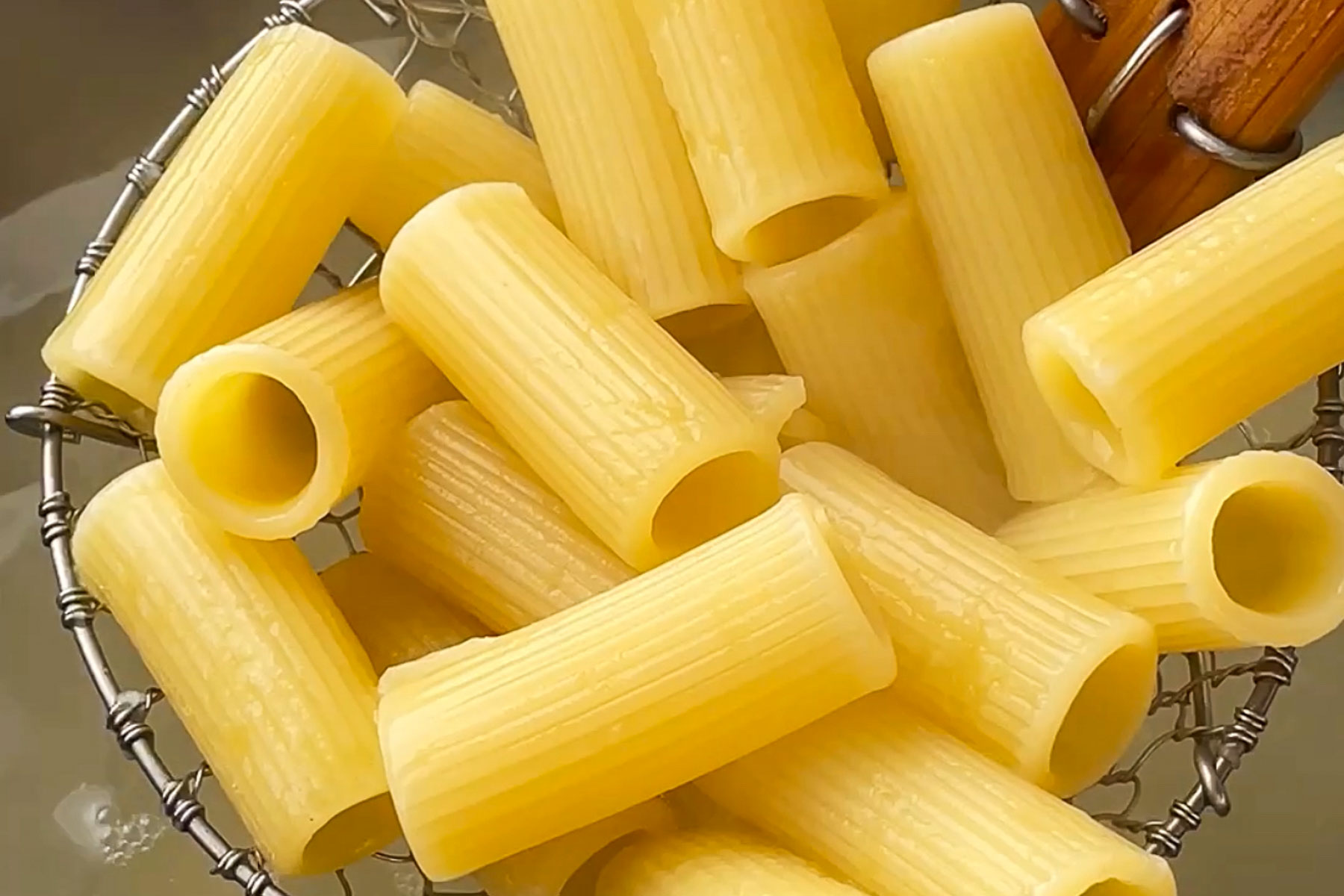Close-up of cooked rigatoni pasta being drained in a metal strainer, with beige, ridged tubes of pasta and some water droplets visible.