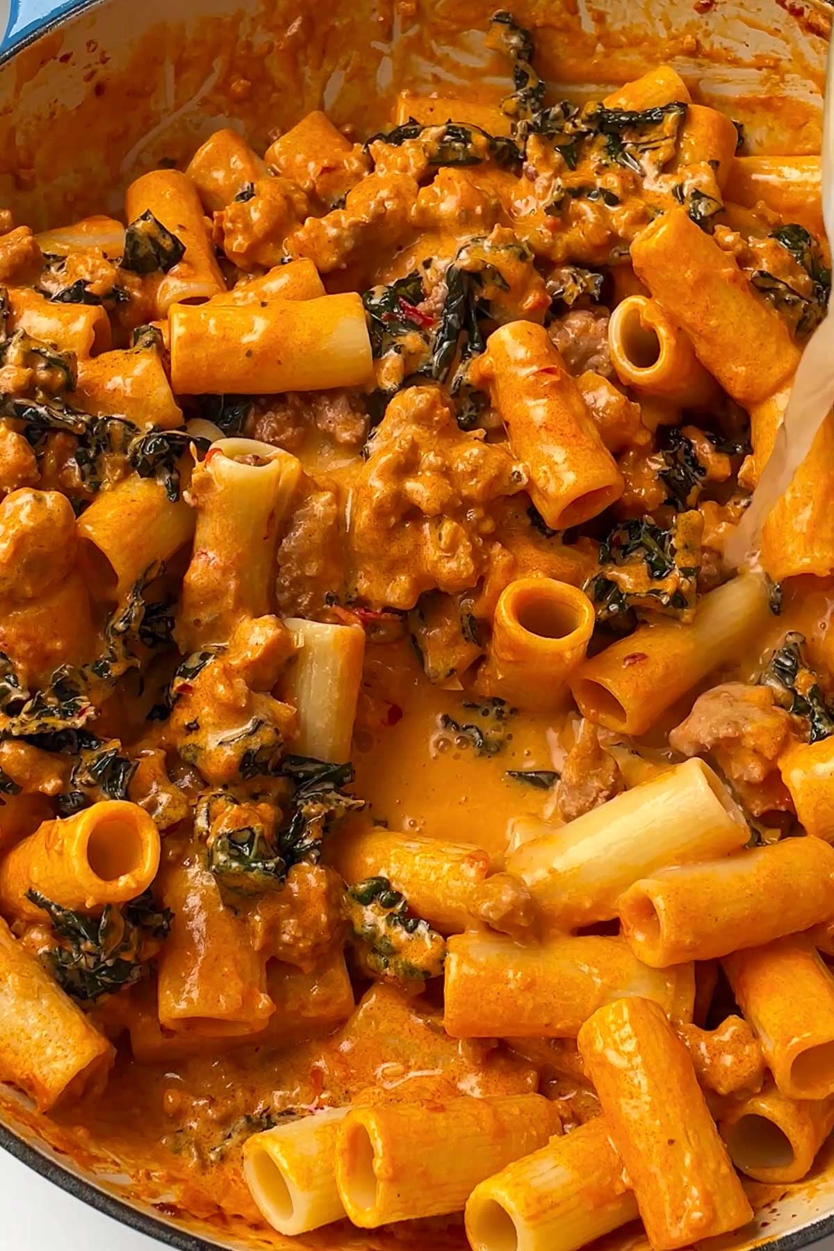 Creamy rigatoni pasta mixed with ground meat, dark leafy greens, and a rich orange tomato sauce in a large pan.