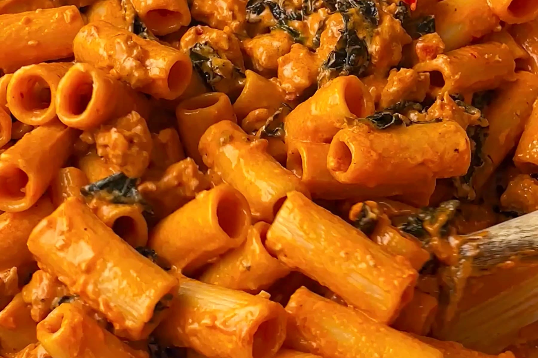 Close-up of cooked rigatoni pasta mixed with a creamy tomato sauce, wilted spinach, and small pieces of meat. The dish has a rich, orange color and a hearty texture.