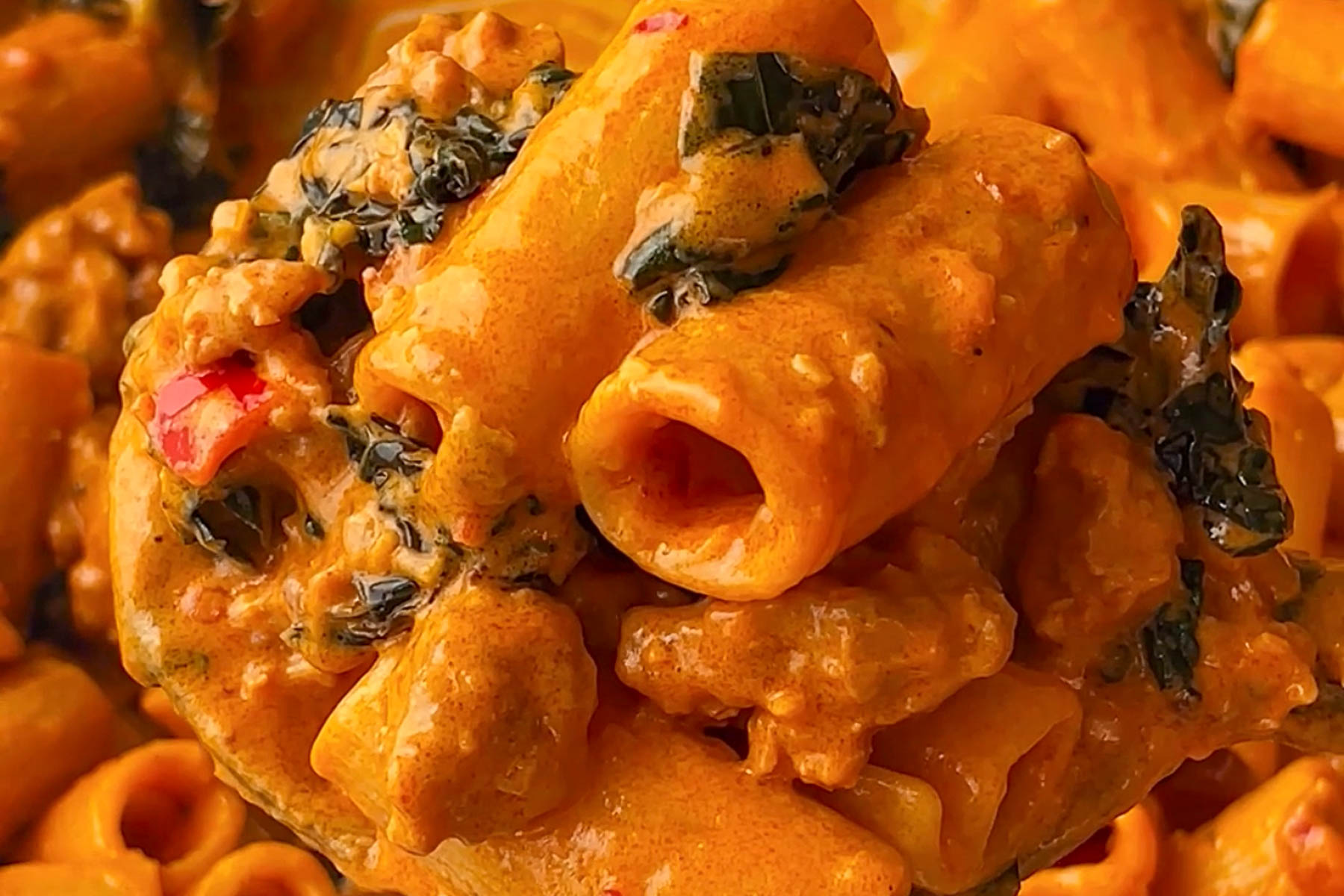 Close-up of creamy rigatoni pasta with sausage, leafy greens, and red bell pepper pieces, coated in a rich orange-colored sauce.