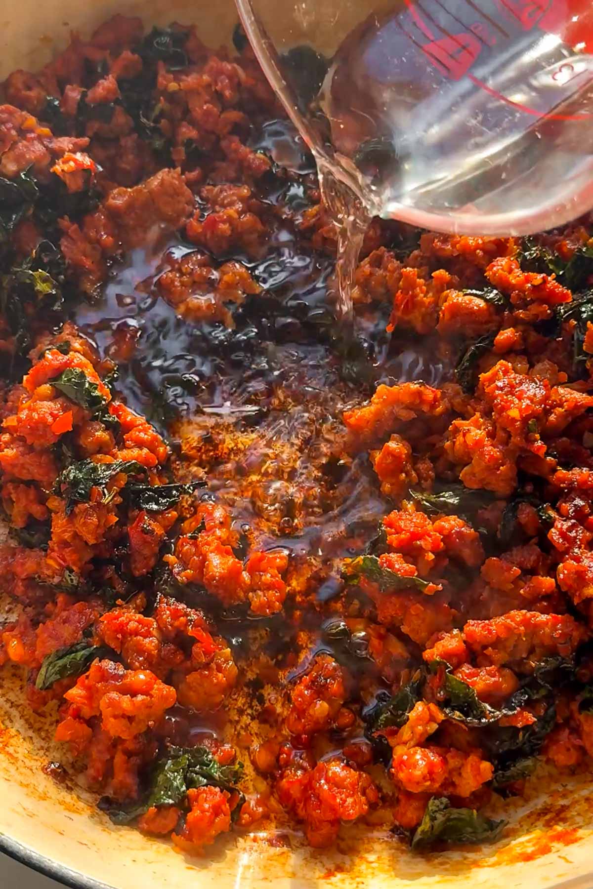 Ground meat with spices and herbs is being cooked in a pan. A clear liquid is being poured into the mixture from a measuring cup, causing it to sizzle and steam as it mixes with the ingredients.