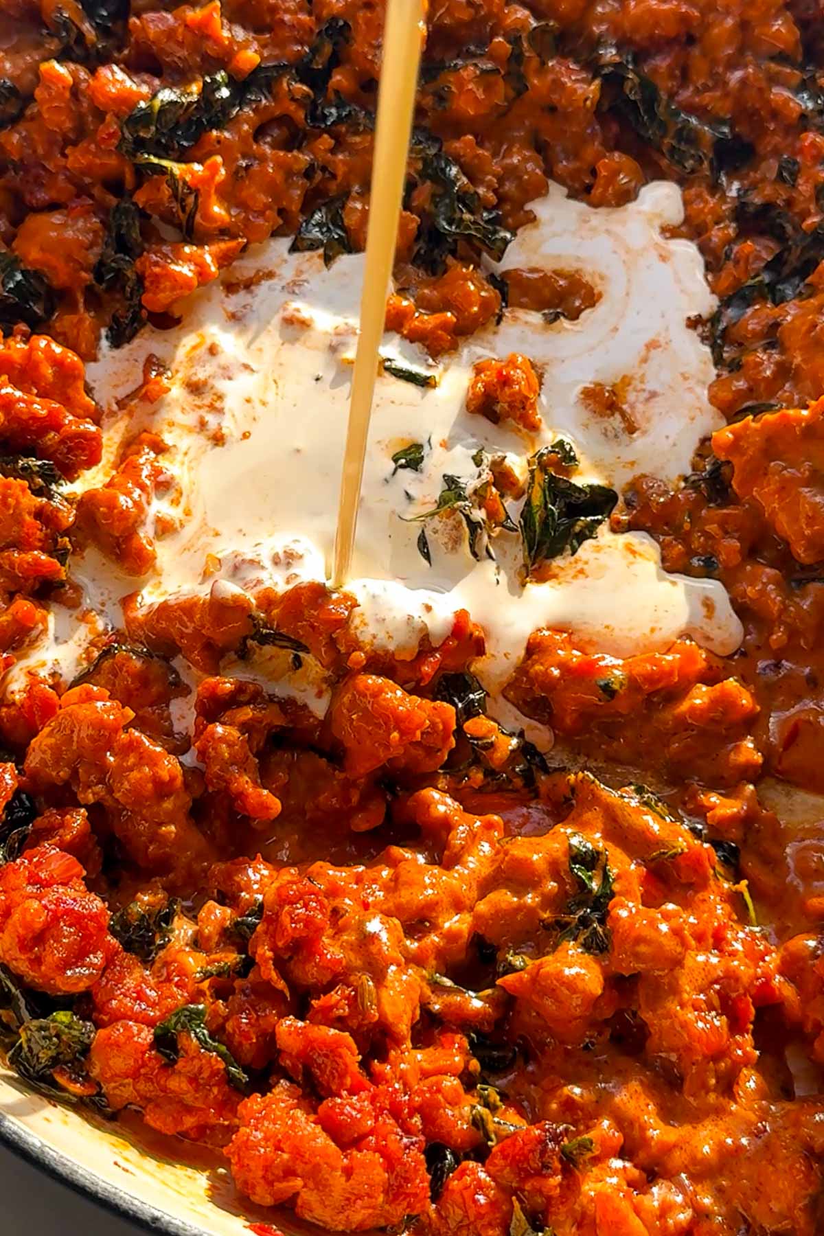 A close-up of a skillet filled with a rich, red tomato and ground meat mixture, spinach, and cream being poured in, creating a creamy swirl in the sauce.