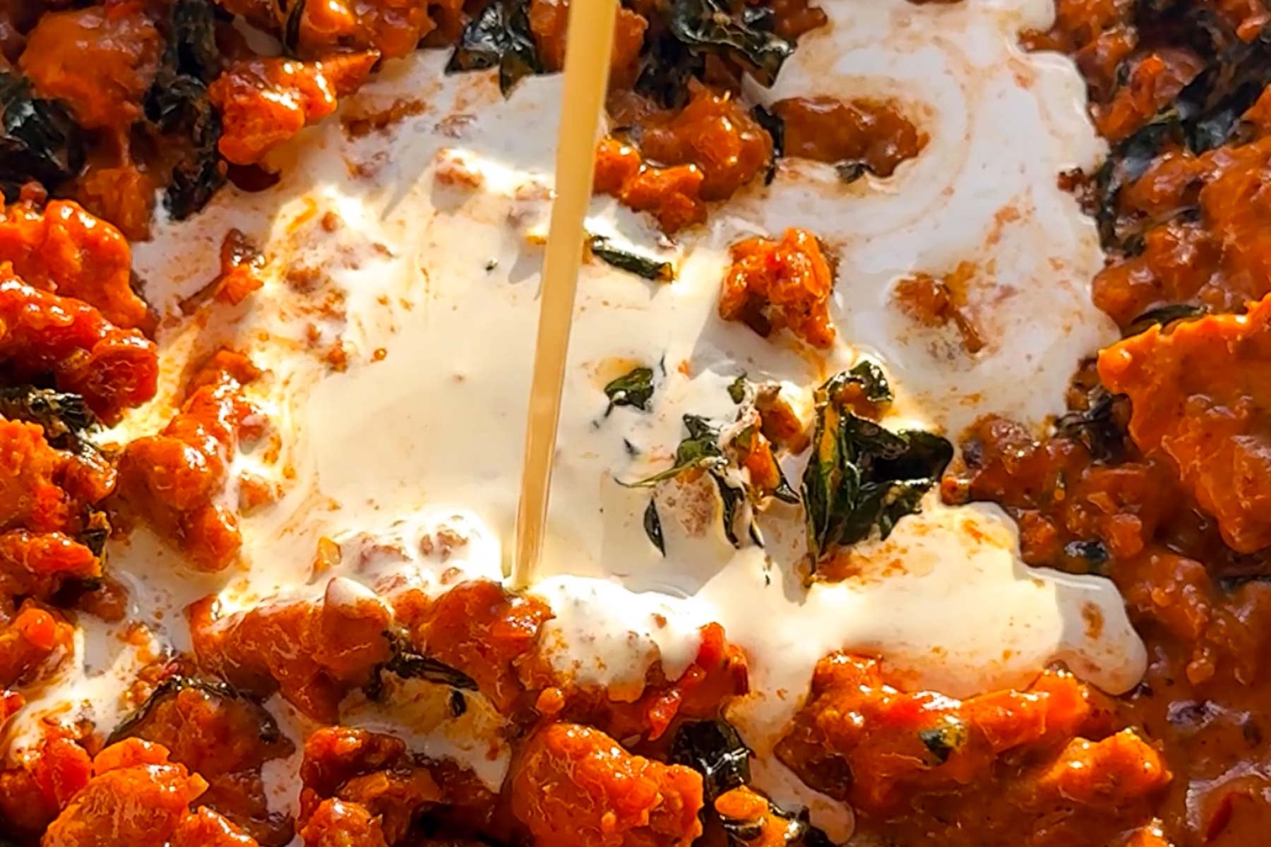 A close-up of a vibrant orange curry with chunks of meat and leafy greens, as a swirl of cream is being poured in, mixing into the rich, saucy dish.