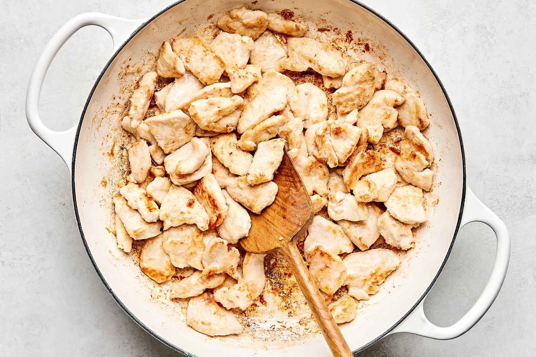 Sautéed pieces of chicken breast are cooking in a large white skillet, with a wooden spoon resting among the cooked chicken pieces.