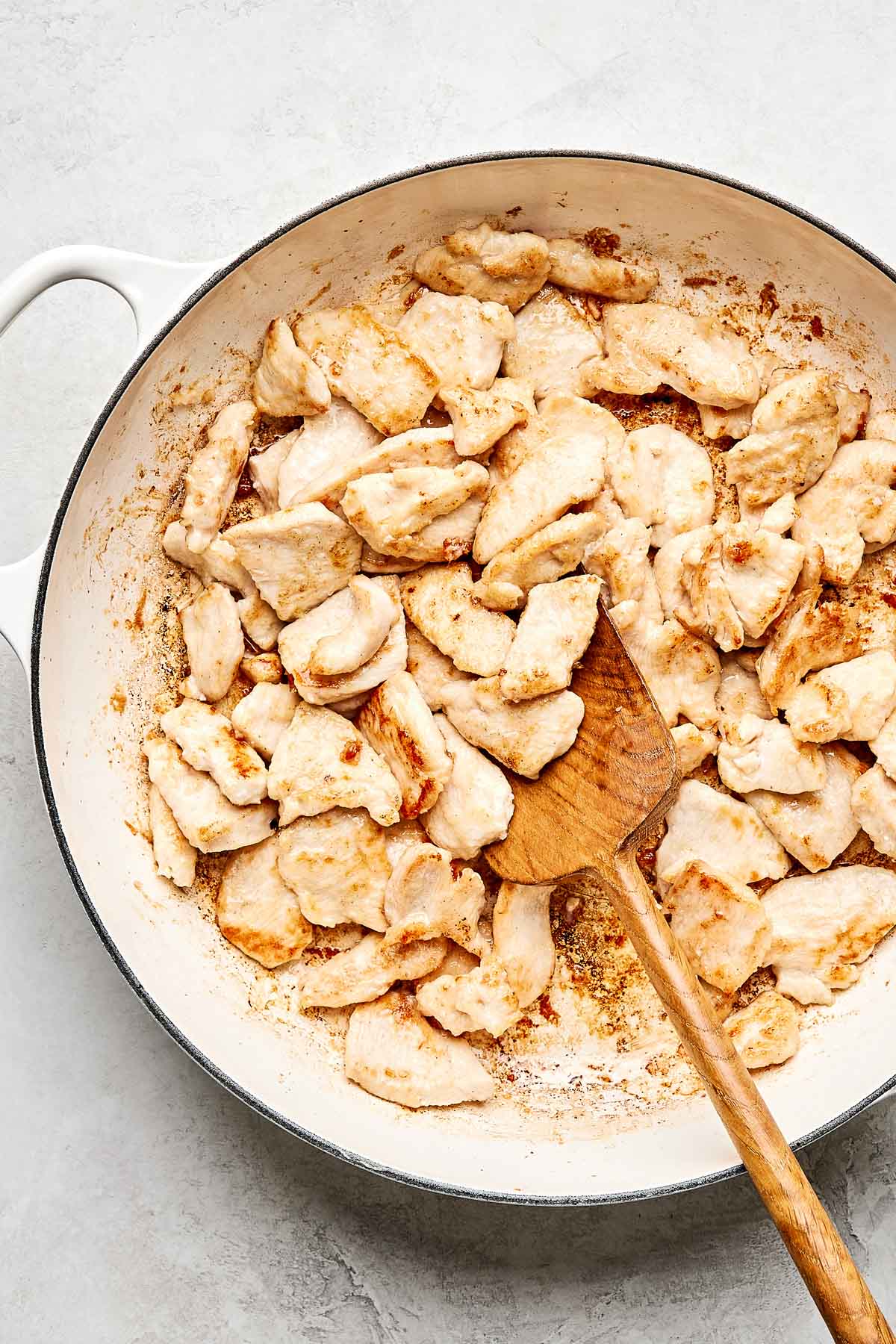 A white skillet filled with cooked, golden-brown chicken pieces being stirred with a wooden spatula. The pan is set on a light-colored surface.