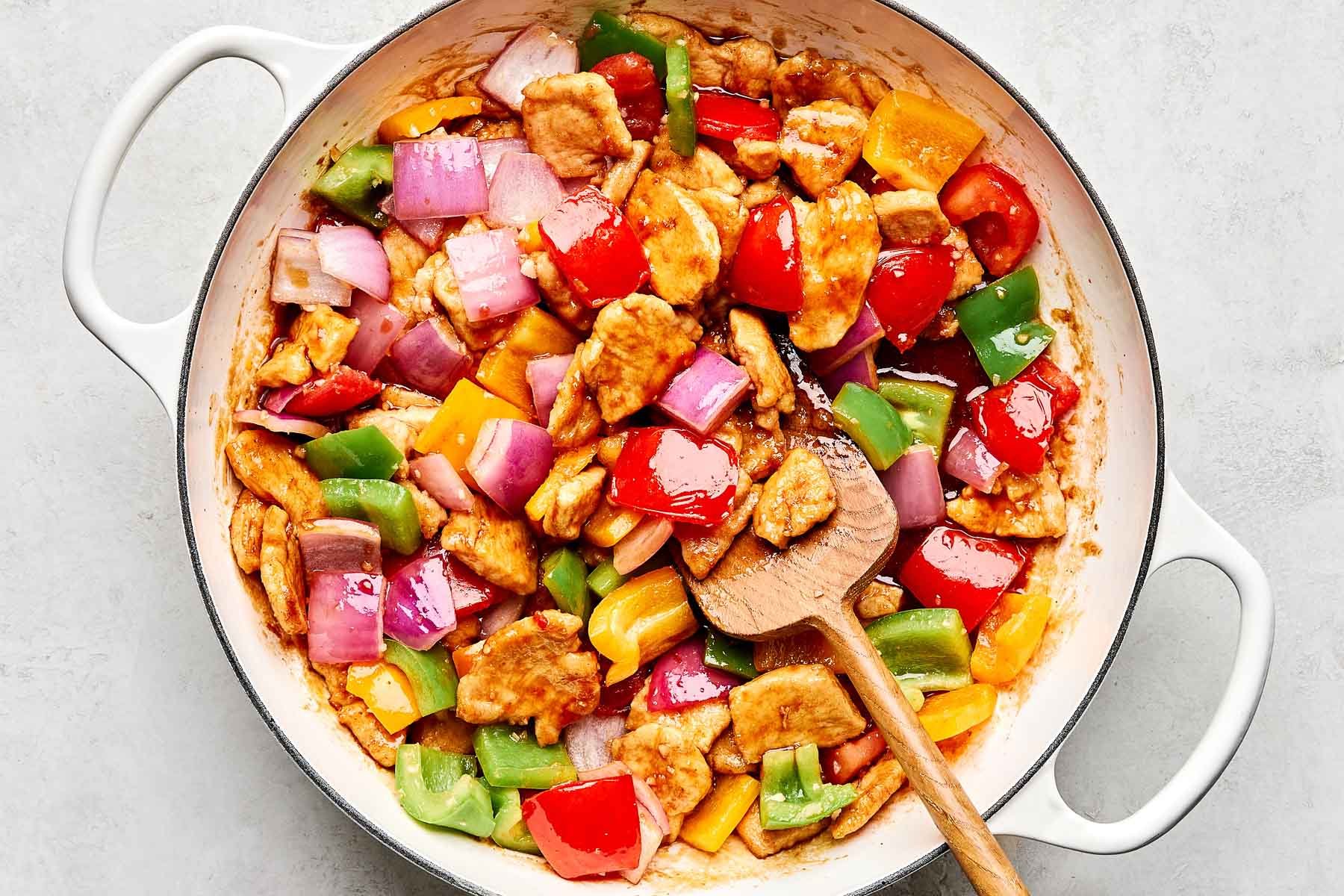 A white pot filled with sweet and sour chicken, featuring pieces of chicken, red onions, and colorful bell peppers, all coated in sauce. A wooden spoon rests inside the pot.