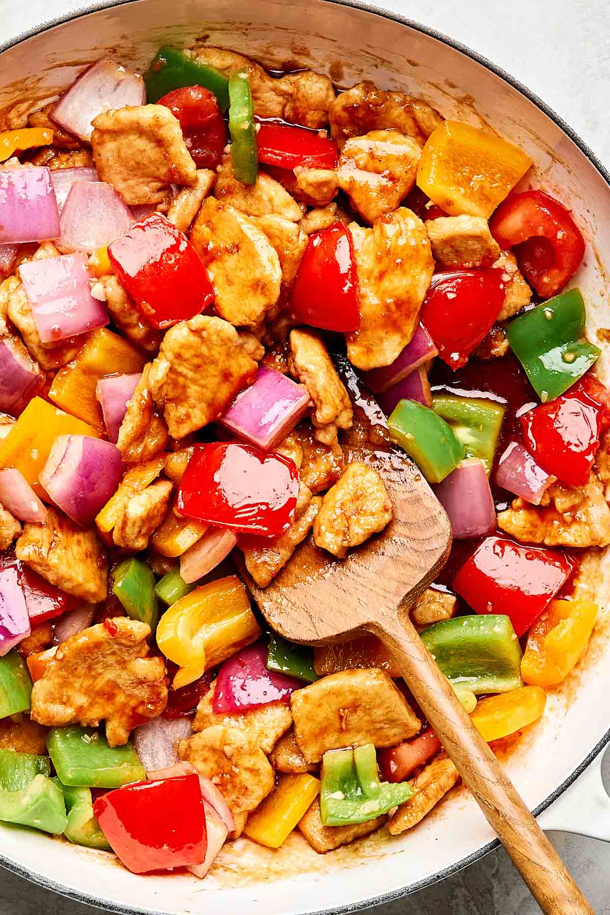 A close-up of a skillet filled with colorful sweet and sour chicken, featuring chunks of chicken, red onions, and green, yellow, and red bell peppers, being stirred with a wooden spoon.